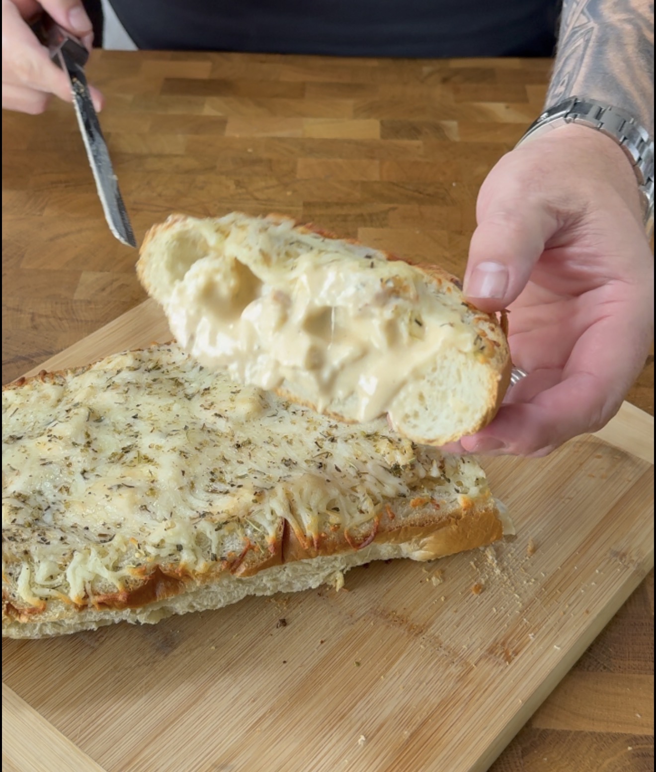 A hand holding a slice of toasted bread topped with melted cheese and herbs, lifted from a larger piece on a wooden cutting board.