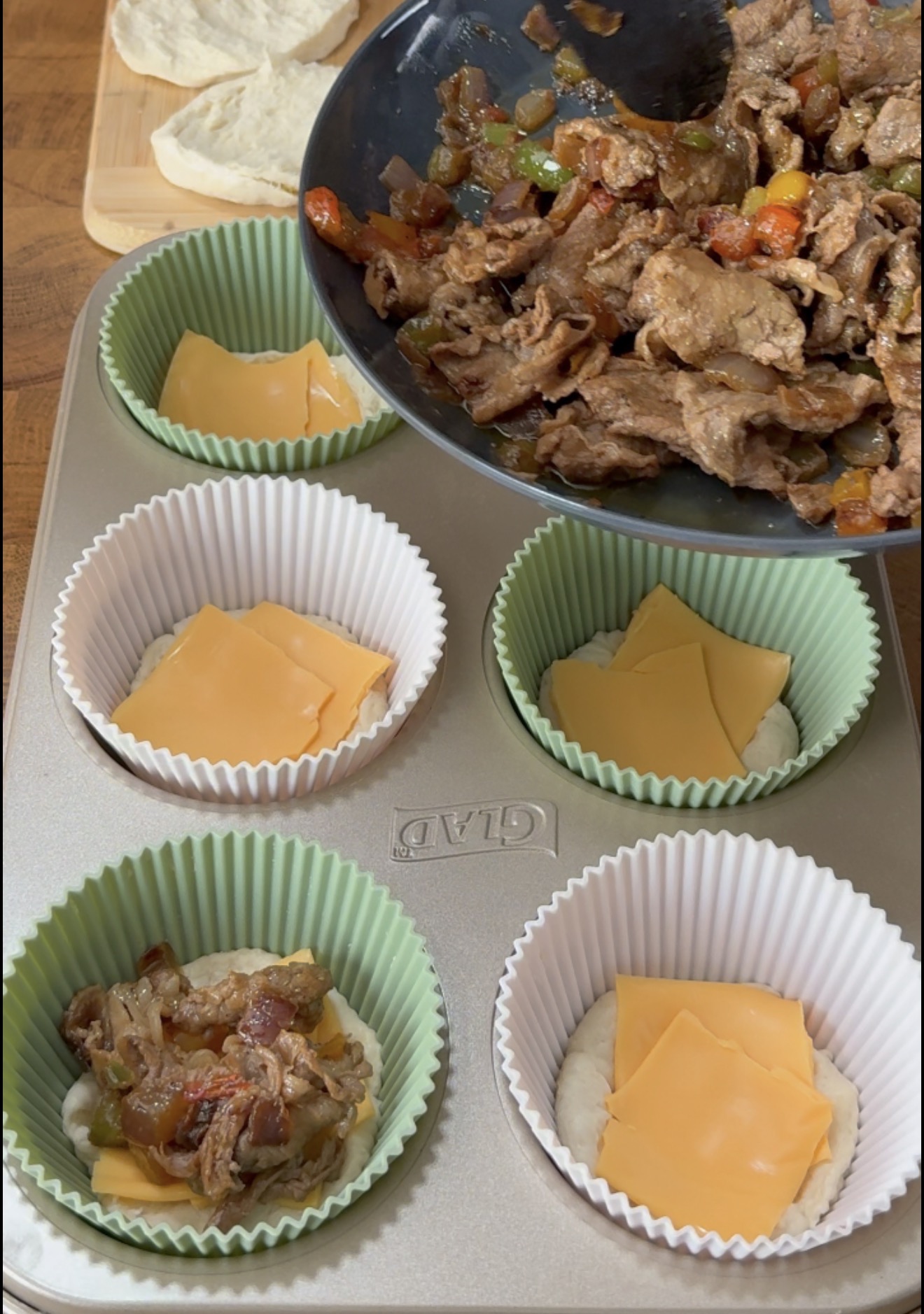A hand uses a spatula to scoop a cooked mixture of thinly sliced beef and peppers into muffin tin cups lined with paper liners and slices of yellow cheese.