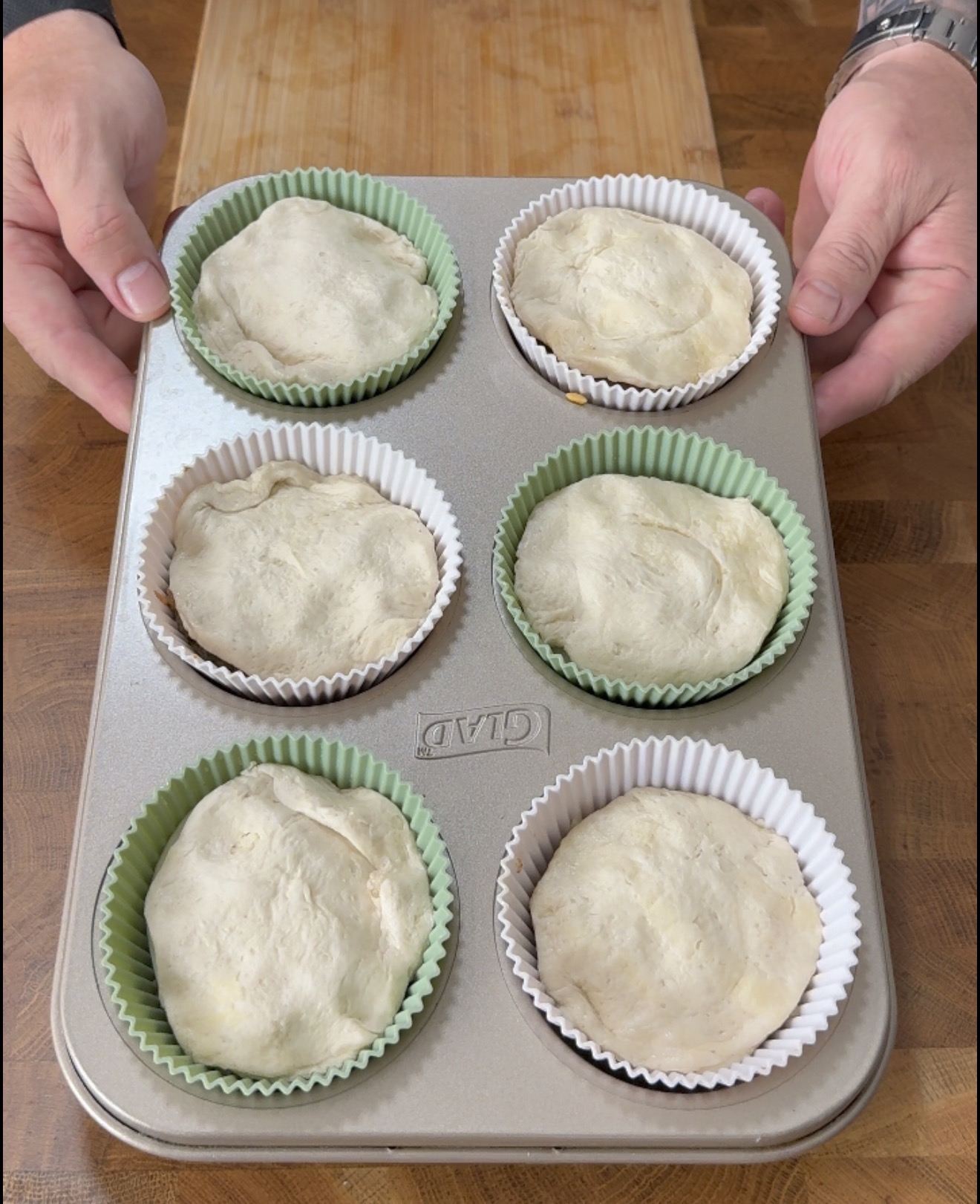 Hands holding a muffin tin containing six portions of raw dough in paper liners.