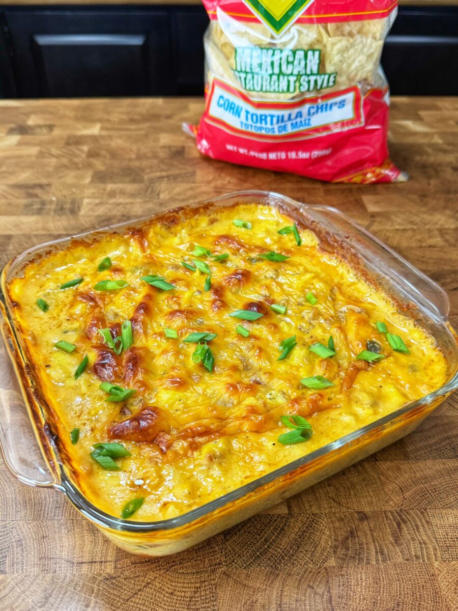 A glass baking dish of cheesy, golden-brown baked dip topped with sliced green onions, with a bag of tortilla chips in the background.