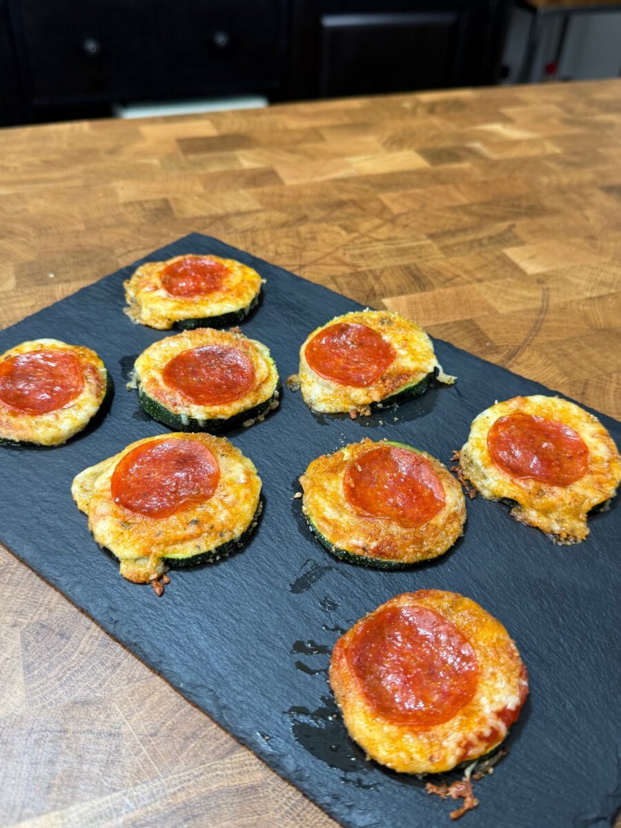 Seven zucchini pizza bites topped with melted cheese and pepperoni on a black slate serving board.