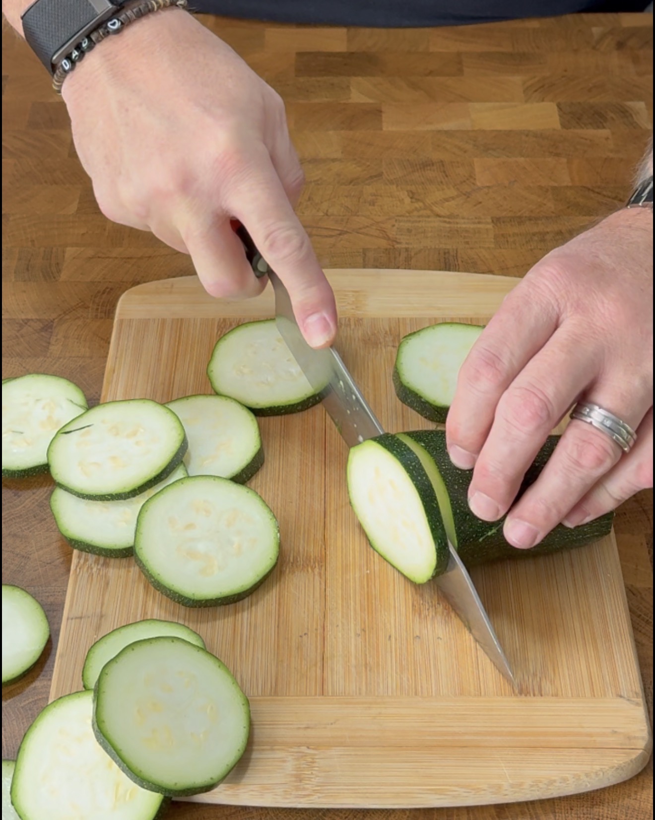 A persons hands using a knife to slice a zucchini into rounds on a wooden cutting board.