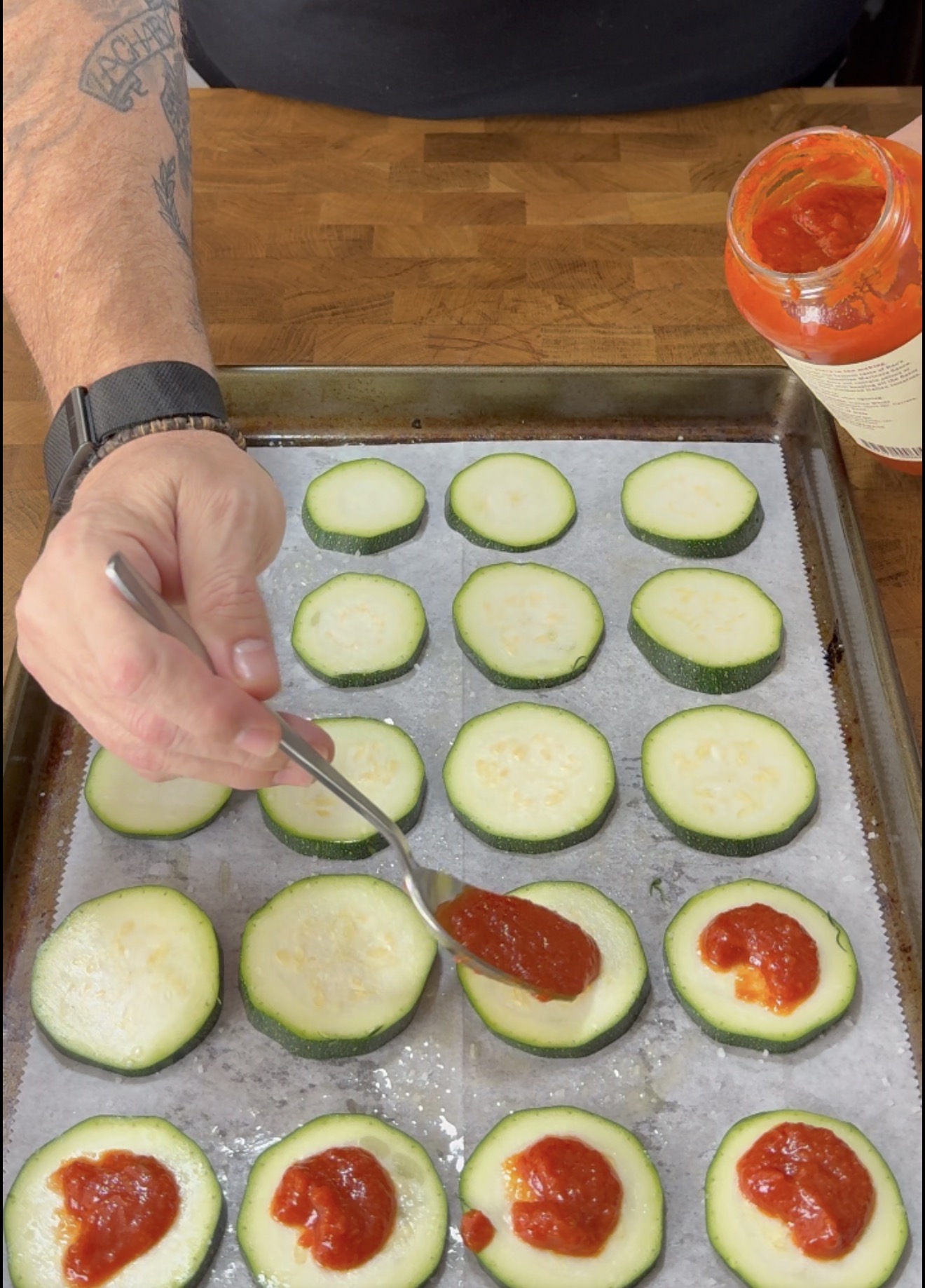 A person uses a spoon to add red sauce onto sliced zucchini rounds arranged on a parchment-lined baking sheet.