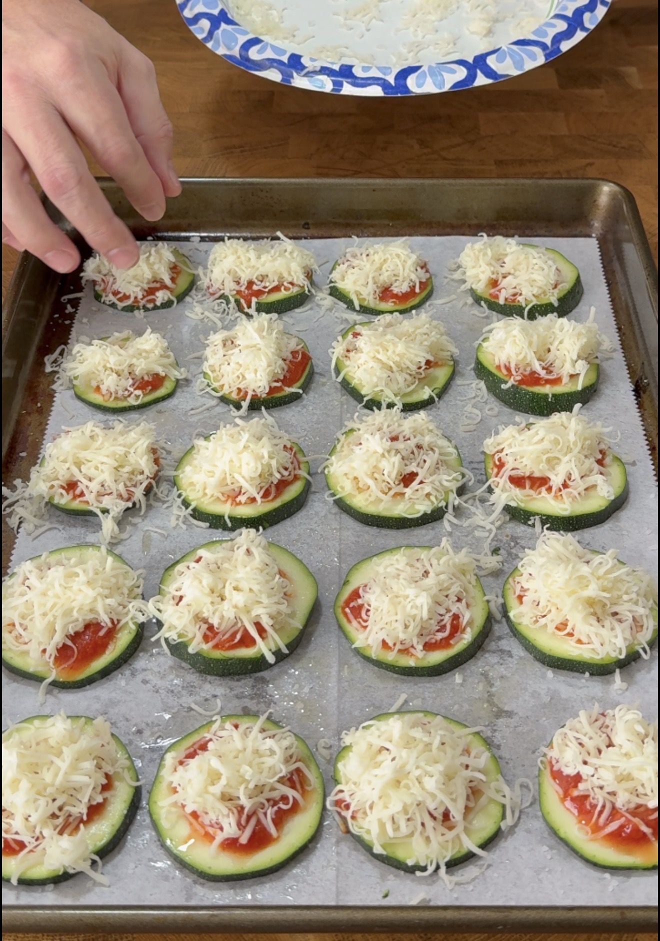 A hand adds shredded cheese to slices of zucchini topped with tomato sauce on a parchment-lined baking sheet.