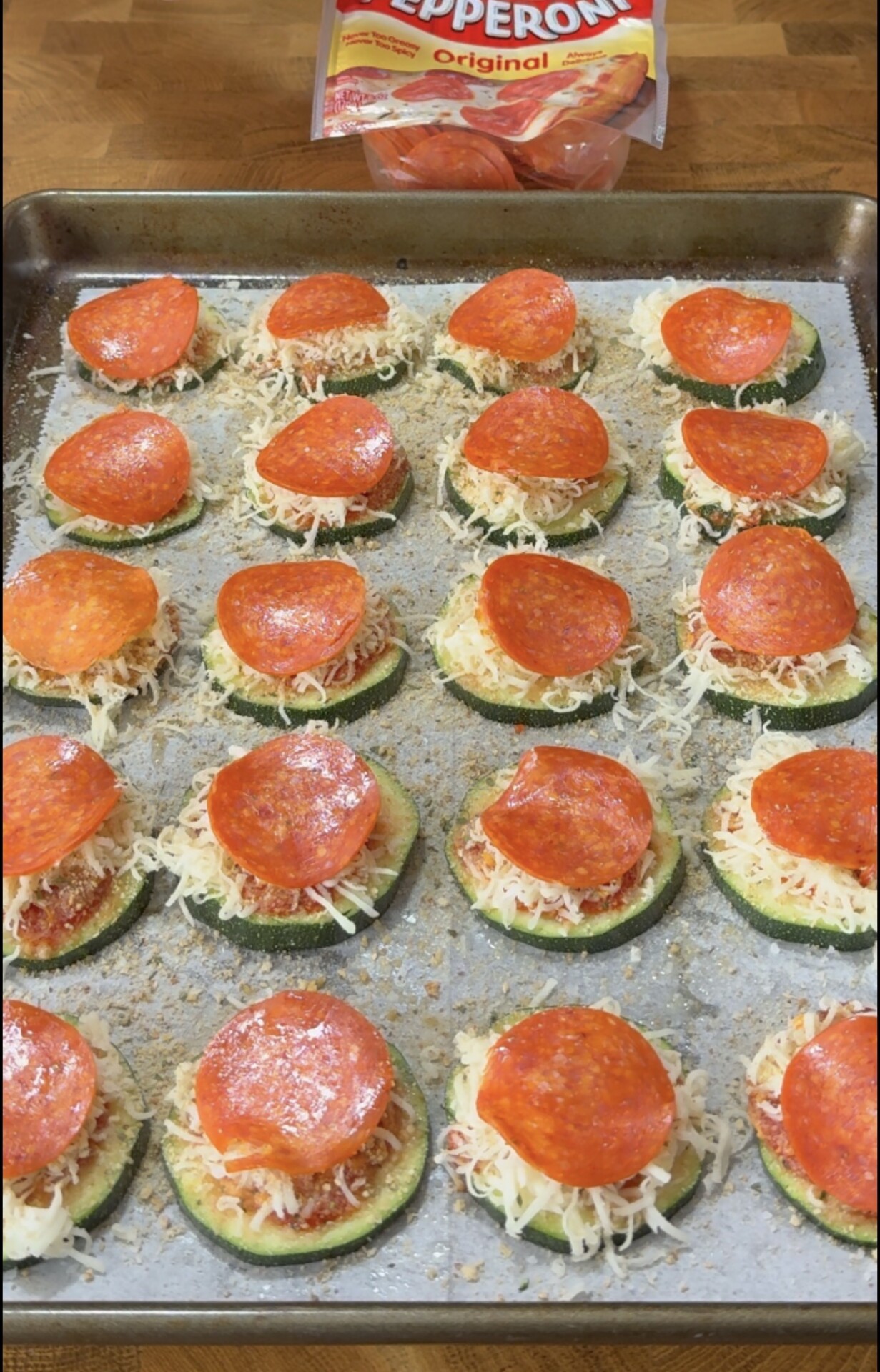 Zucchini slices topped with tomato sauce, shredded cheese, and pepperoni on a parchment-lined baking sheet, with a package of pepperoni above.