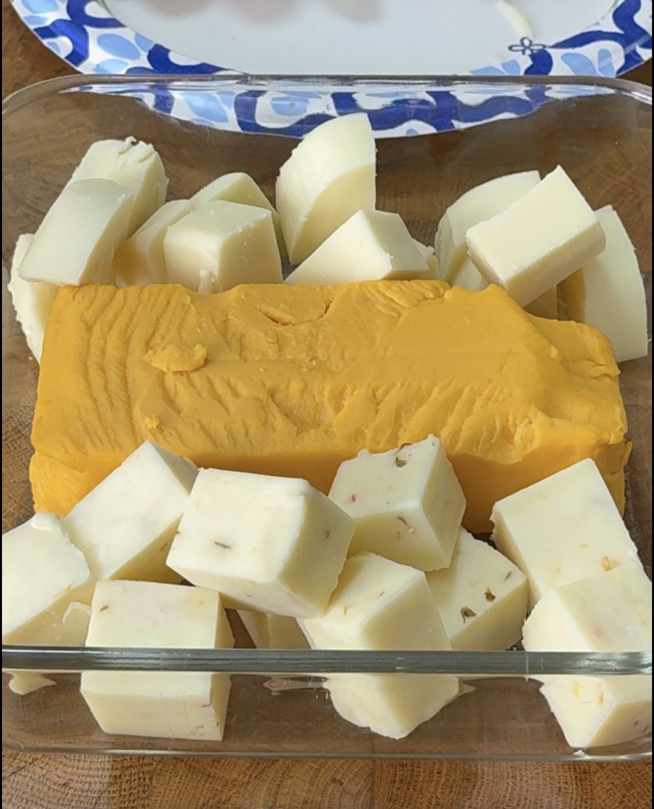 A glass dish filled with a large block of orange cheese surrounded by cubes of white cheese with herbs.