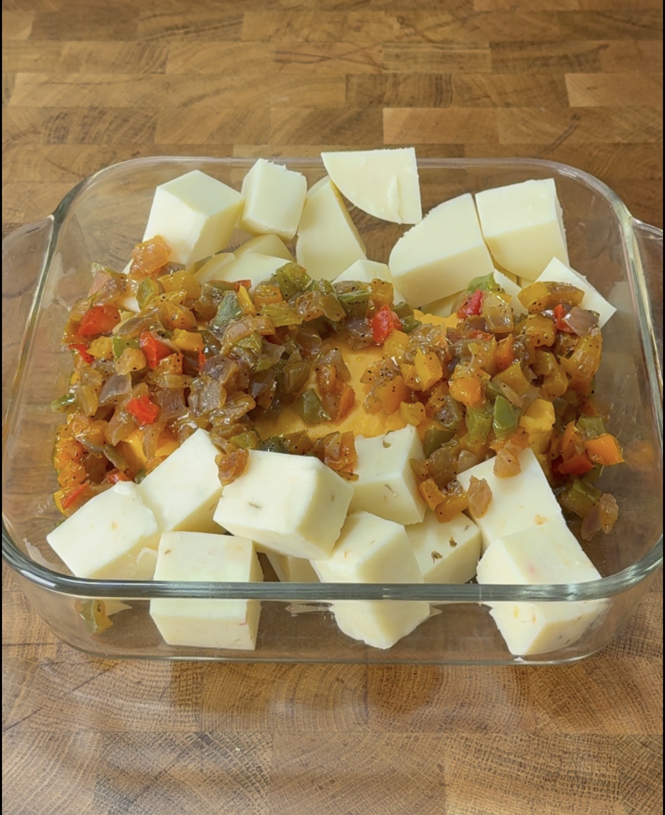 Cubes of white cheese topped with a sautéed mixture of colorful bell peppers and onions in a glass baking dish.