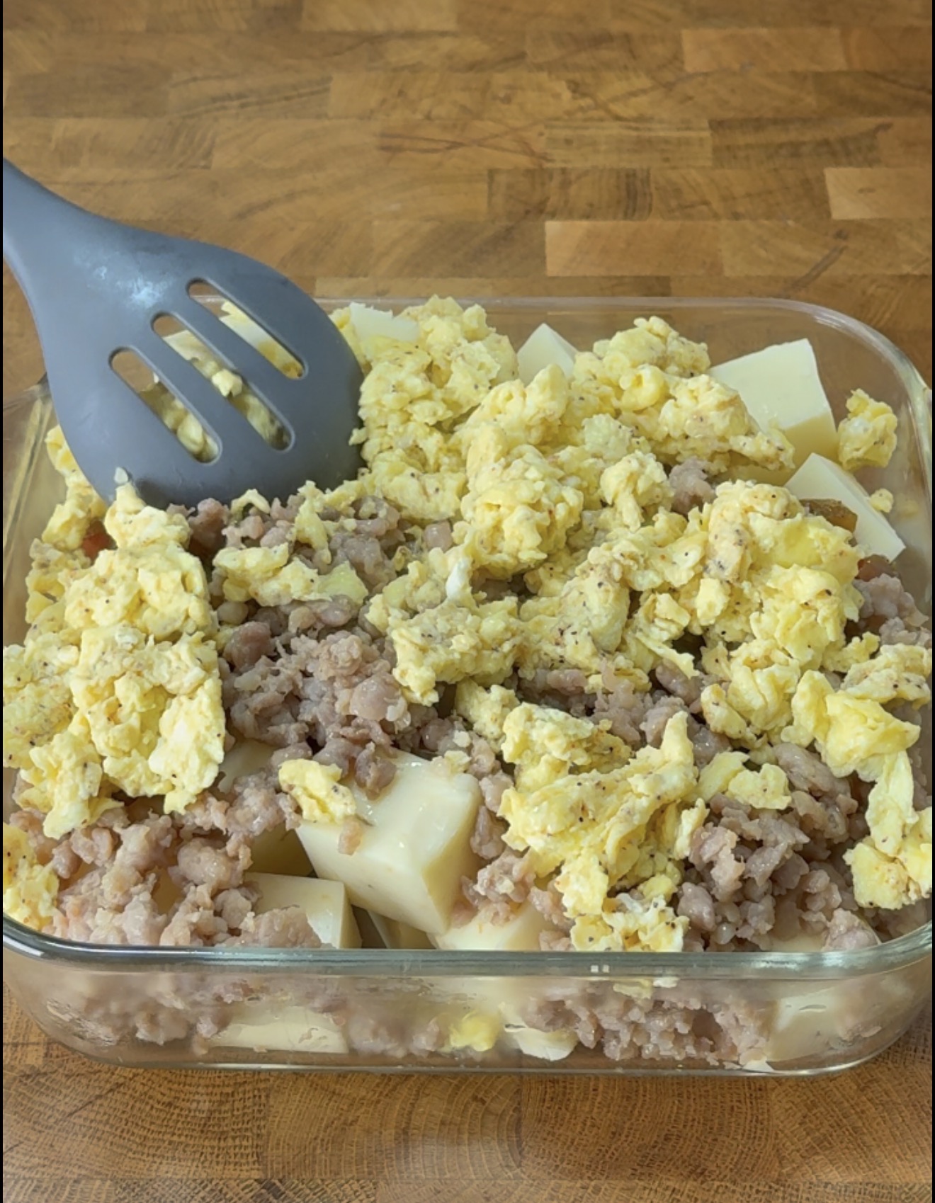 A grey spatula mixes a casserole of cubed cheese, browned sausage, and scrambled eggs in a glass baking dish.