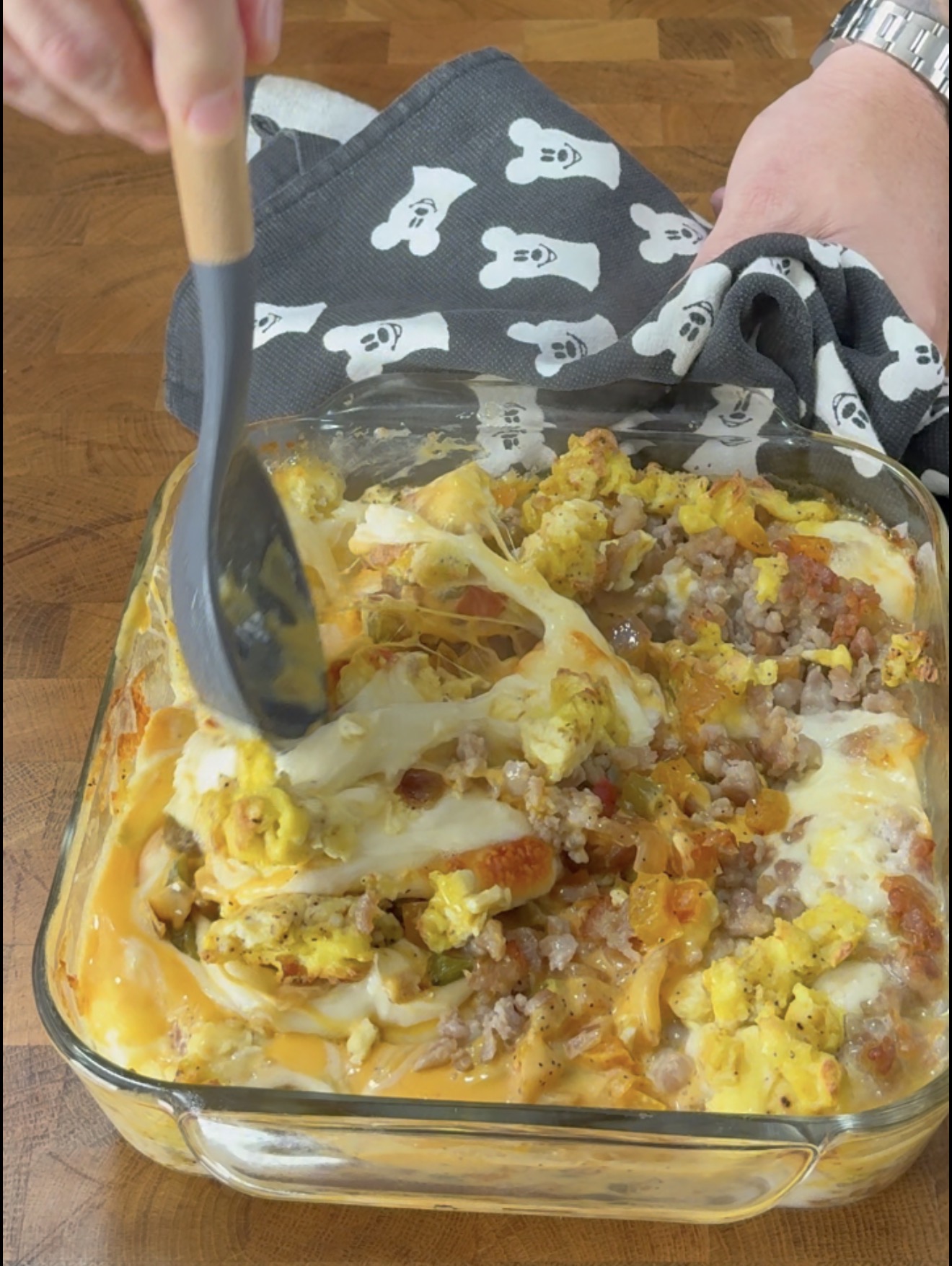 A hand uses a grey spoon to lift a portion of cheesy breakfast casserole containing eggs, sausage, and vegetables from a glass baking dish.