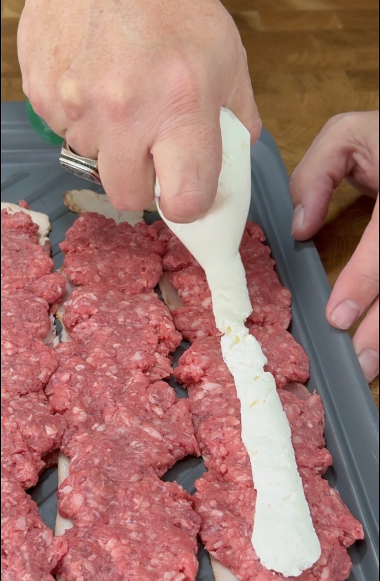 A hand squeezes a log of white cheese onto a tray of raw ground meat patties.