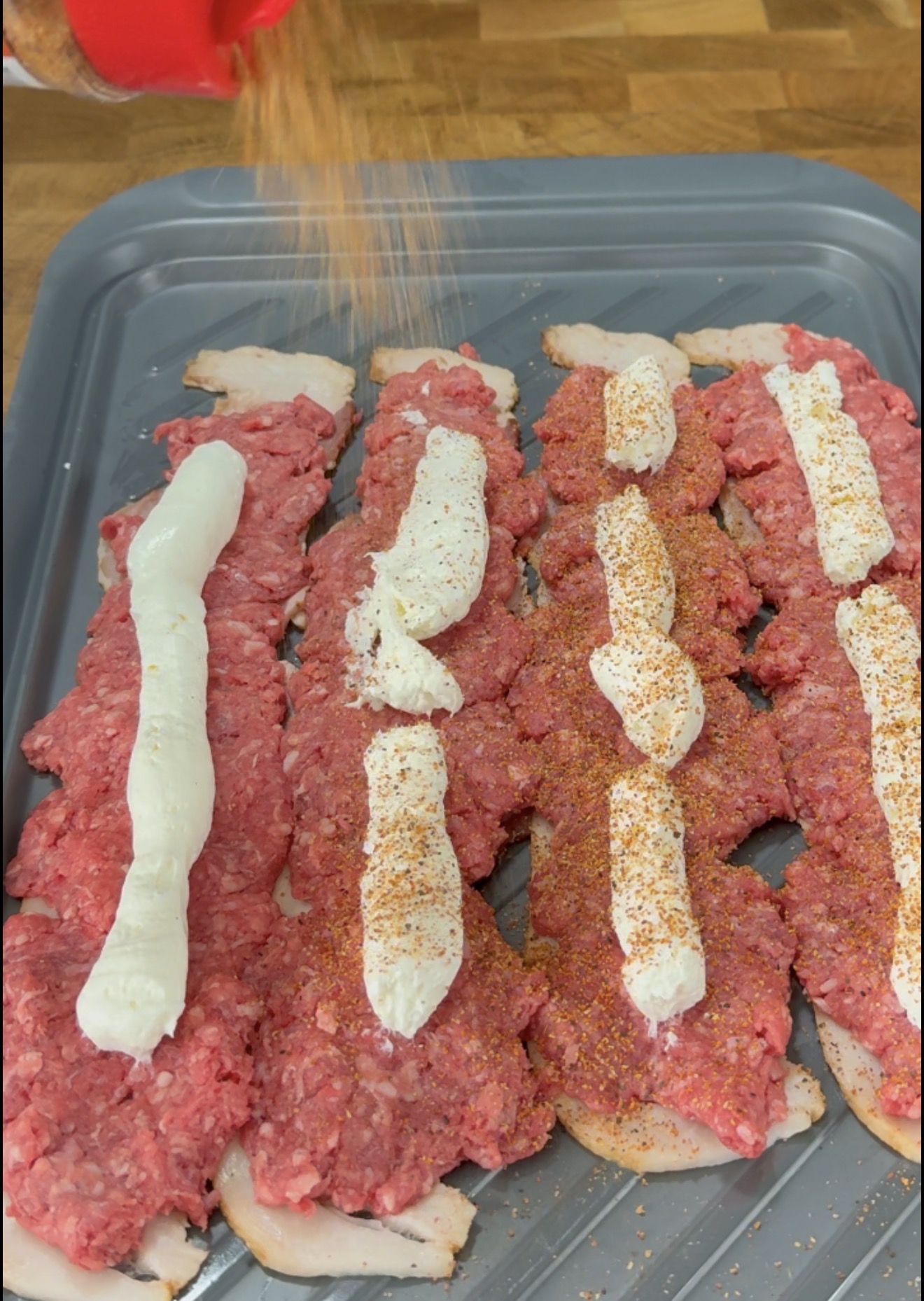 Strips of raw bacon topped with ground meat and smears of cream cheese are being sprinkled with seasoning on a baking sheet.