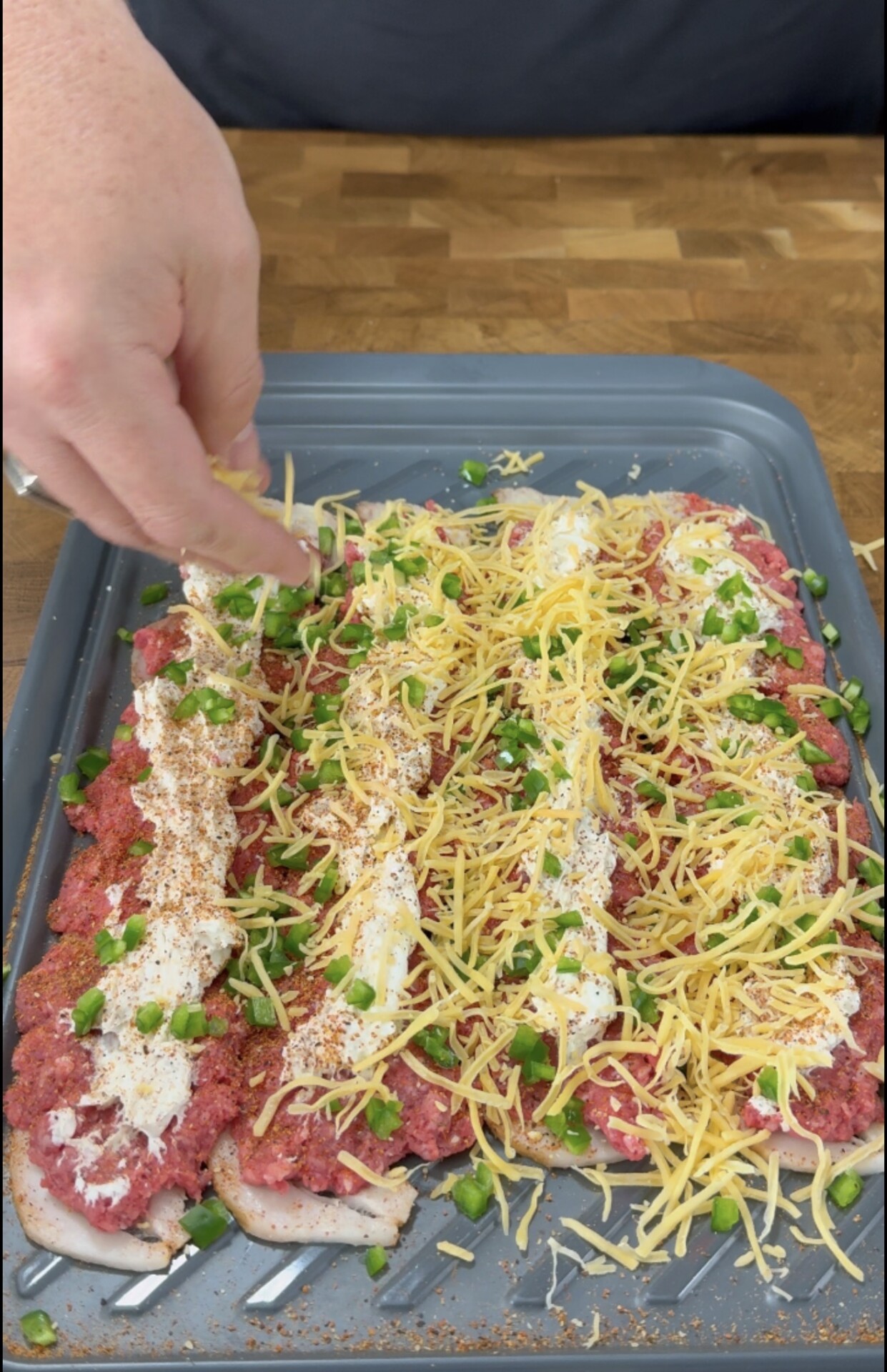 A hand sprinkles shredded cheese onto a baking sheet layered with strips of raw meat topped with dollops of white cream, spices, and chopped green peppers.