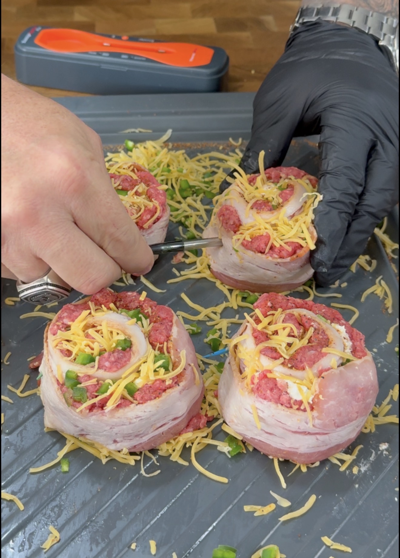 Hands in black gloves place metal skewers into bacon-wrapped meat rolls topped with shredded cheese and chopped green peppers.