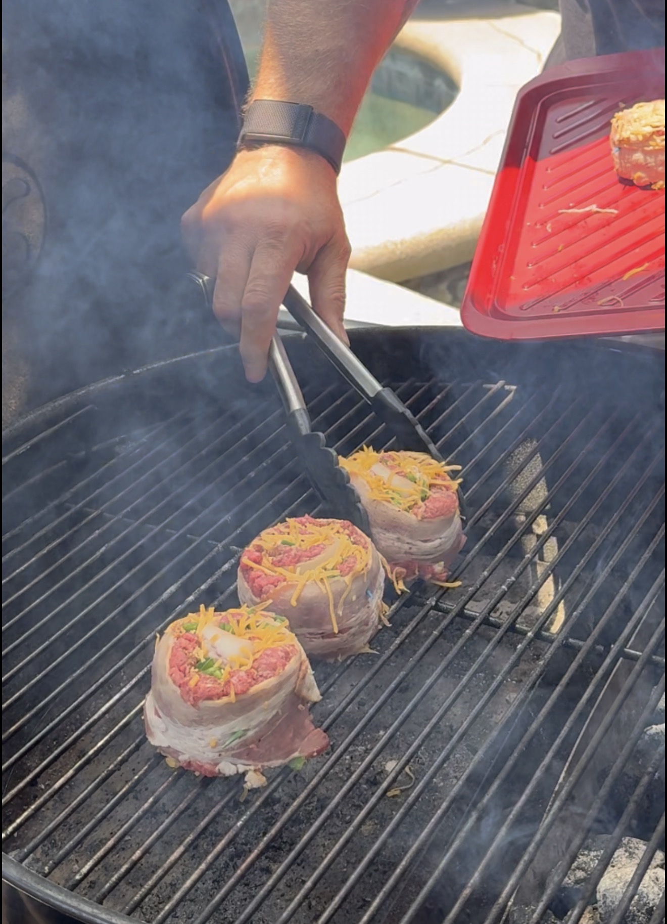 A person using tongs to place bacon-wrapped, cheese-topped burger patties onto a hot grill.