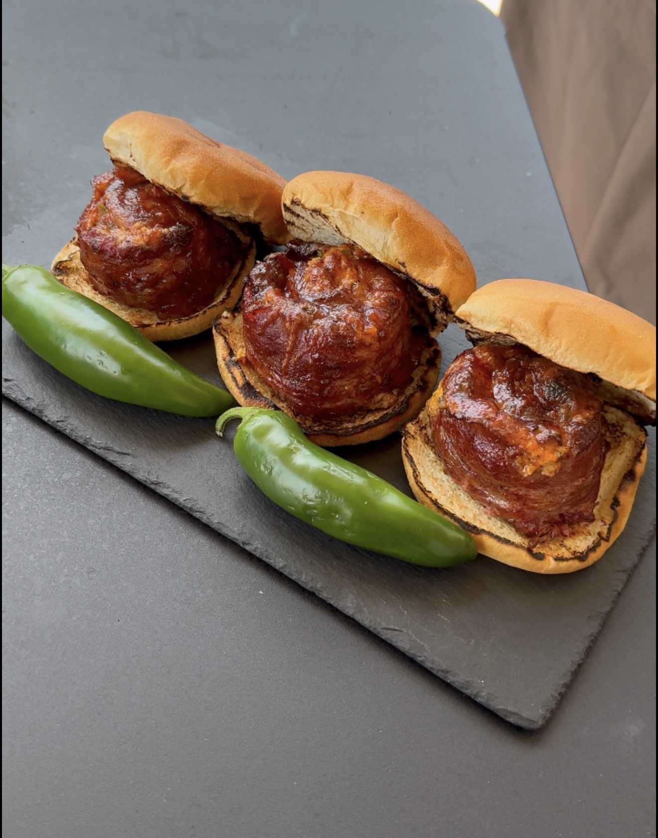 Three meatball sliders with sauce on toasted buns, served on a slate board with two whole jalapeño peppers.
