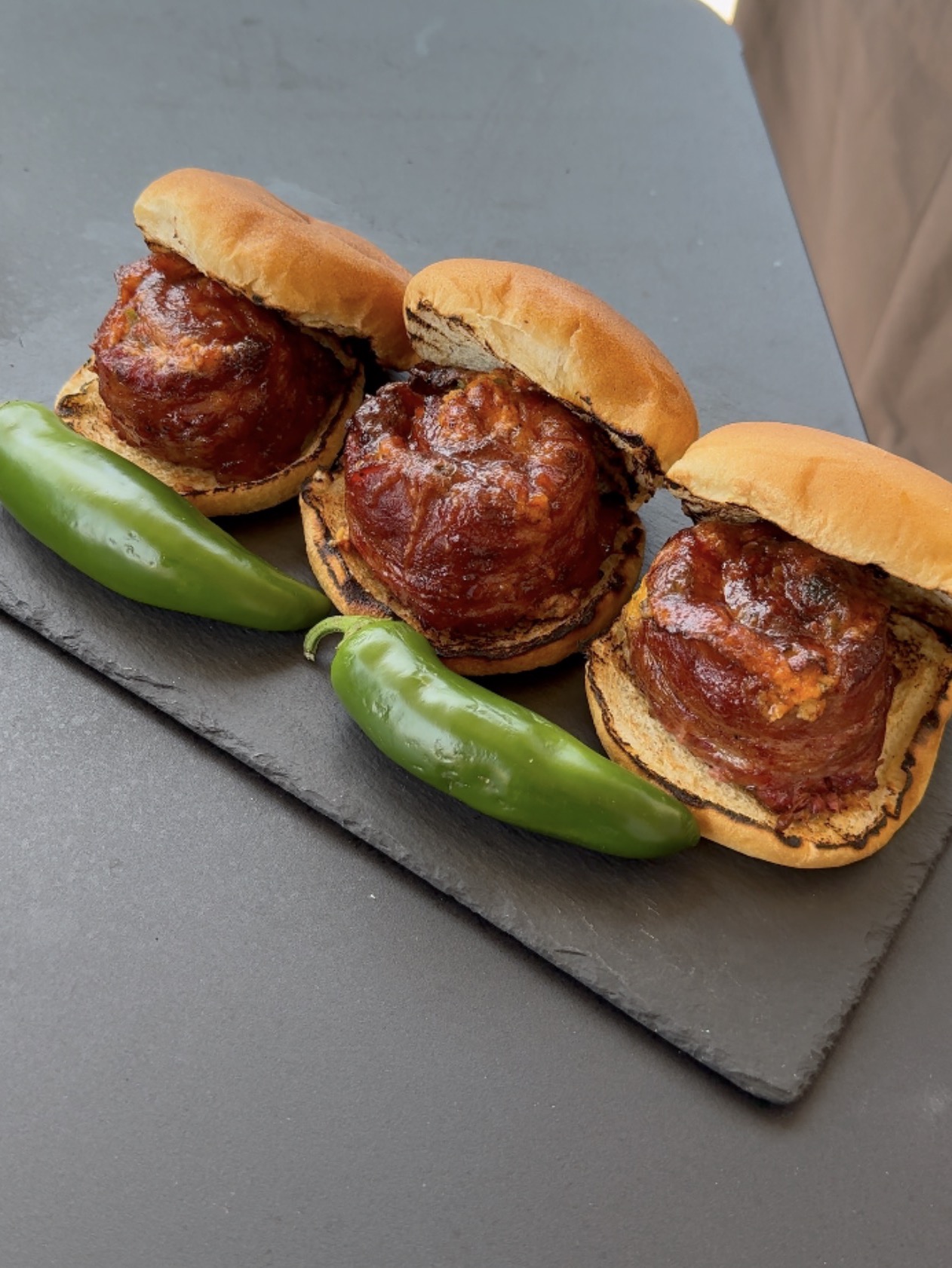 Three meatball sliders with barbecue sauce on buns, served on a slate board with two whole green chili peppers.