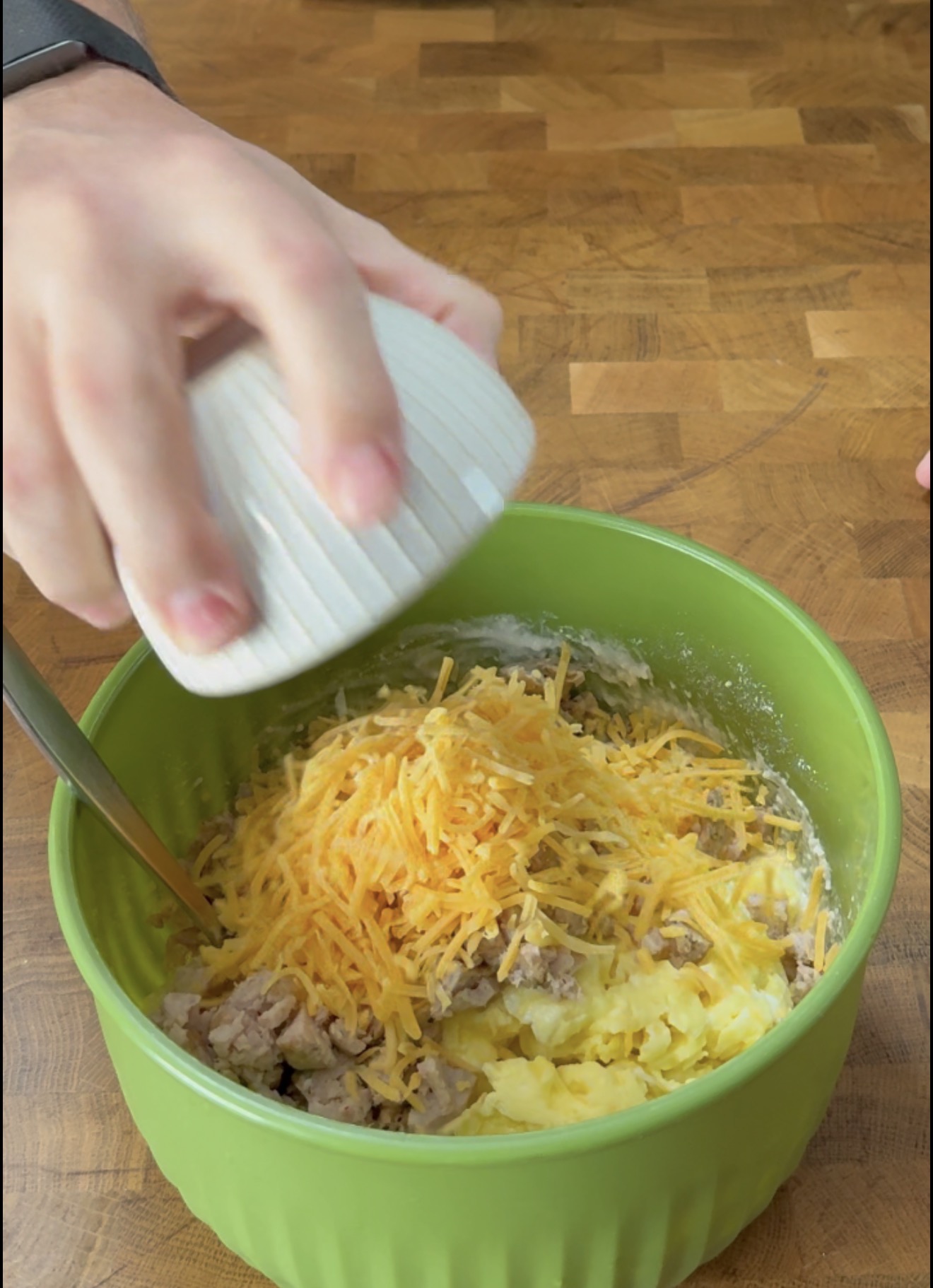 A hand holds a small white bowl over a green mixing bowl filled with crumbled sausage, scrambled eggs, and shredded cheddar cheese.