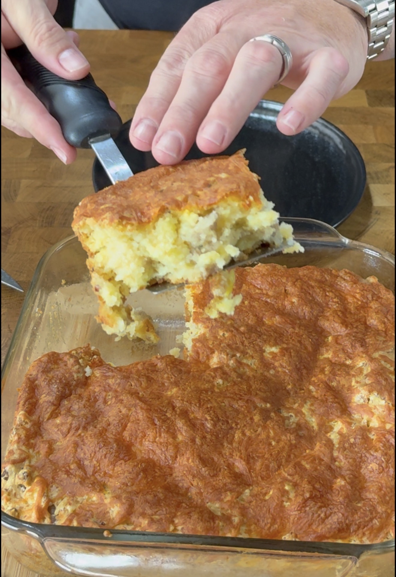 A person uses a spatula to lift a square serving of golden-brown casserole from a glass baking dish.