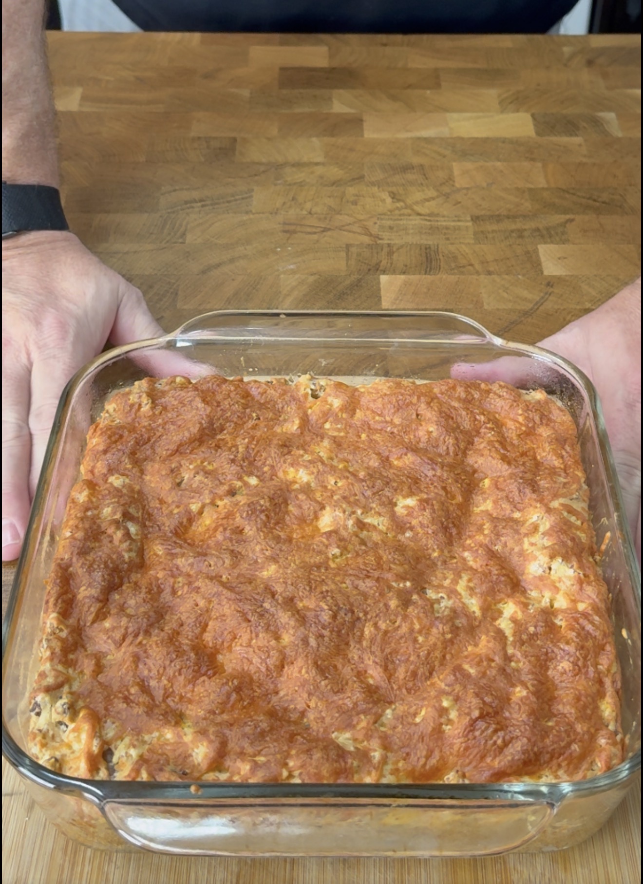 A square glass baking dish filled with a baked casserole topped with melted, browned cheese, held by two hands on a wooden countertop.
