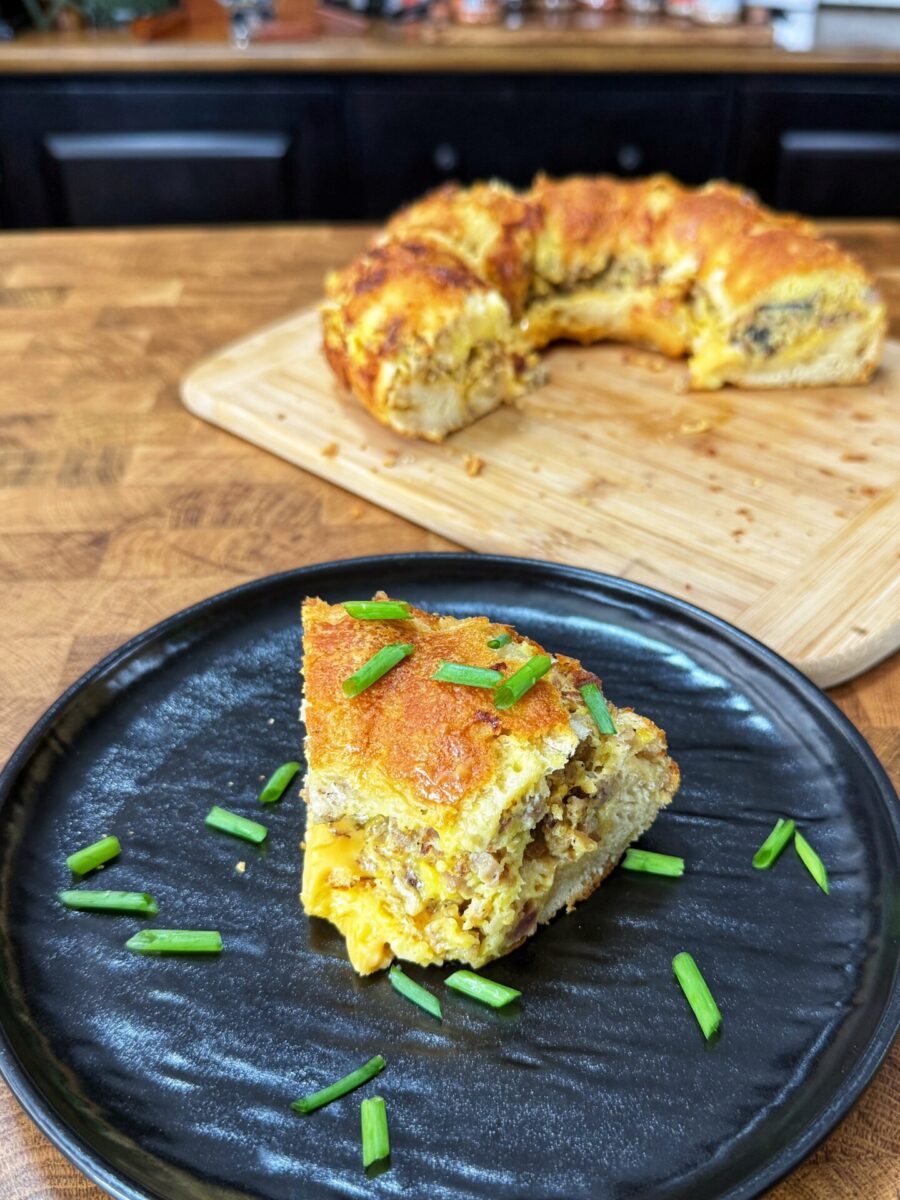 A slice of savory breakfast ring with eggs, cheese, and sausage, garnished with green onions on a black plate, with the remaining ring on a wooden cutting board in the background.