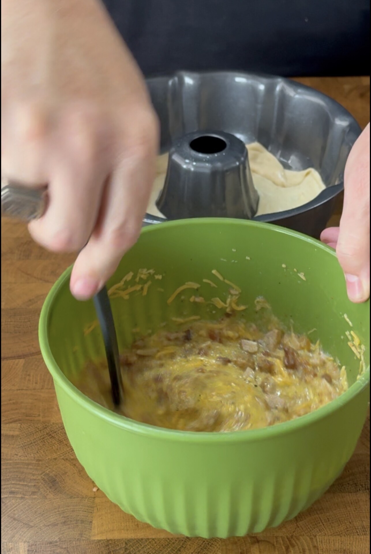 Hands mixing ingredients in a green bowl, with a bundt pan containing dough in the background.
