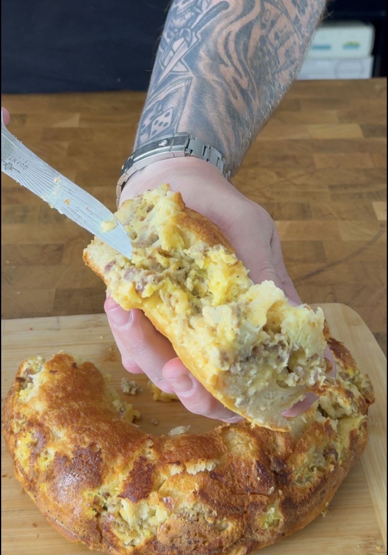 A hand holds a slice of a golden-brown sausage and cheese ring, while a knife rests against the cut side of the pastry.