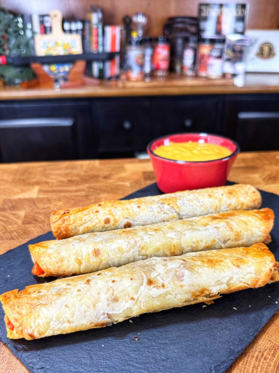 Three rolled flour tortillas on a dark slate board with a red bowl of cheese dip.