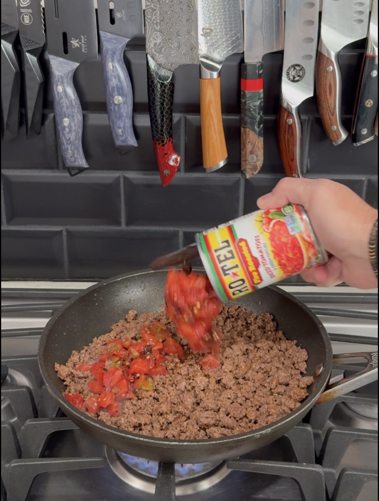 A hand pours a can of Ro-Tel diced tomatoes and green chilies into a pan of browned ground meat on a stovetop.