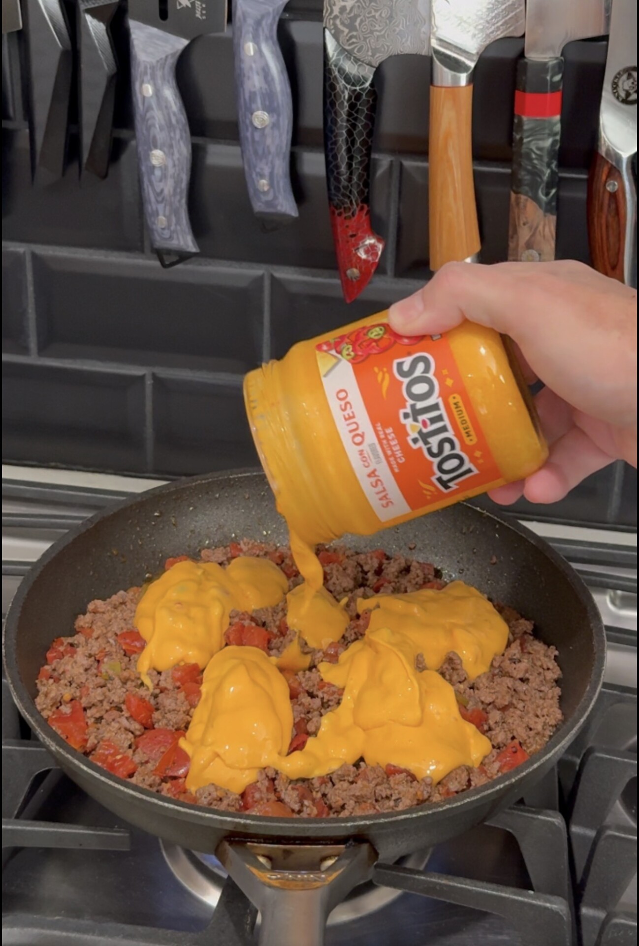 A hand pours a jar of Tostitos salsa con queso onto ground beef and diced tomatoes cooking in a skillet.