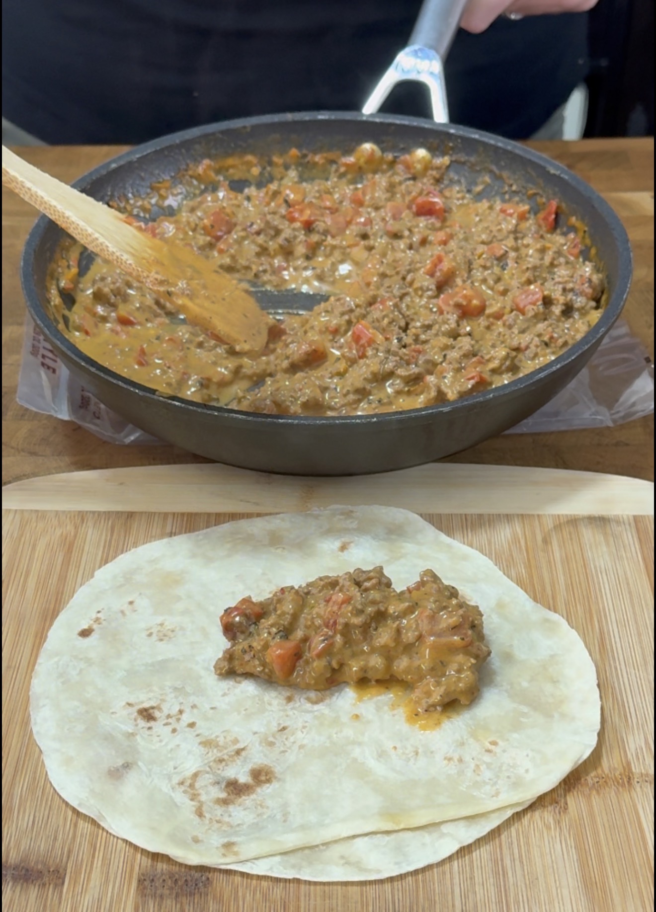 A spoonful of creamy, tomato-based ground meat mixture being placed onto a flour tortilla on a wooden cutting board.