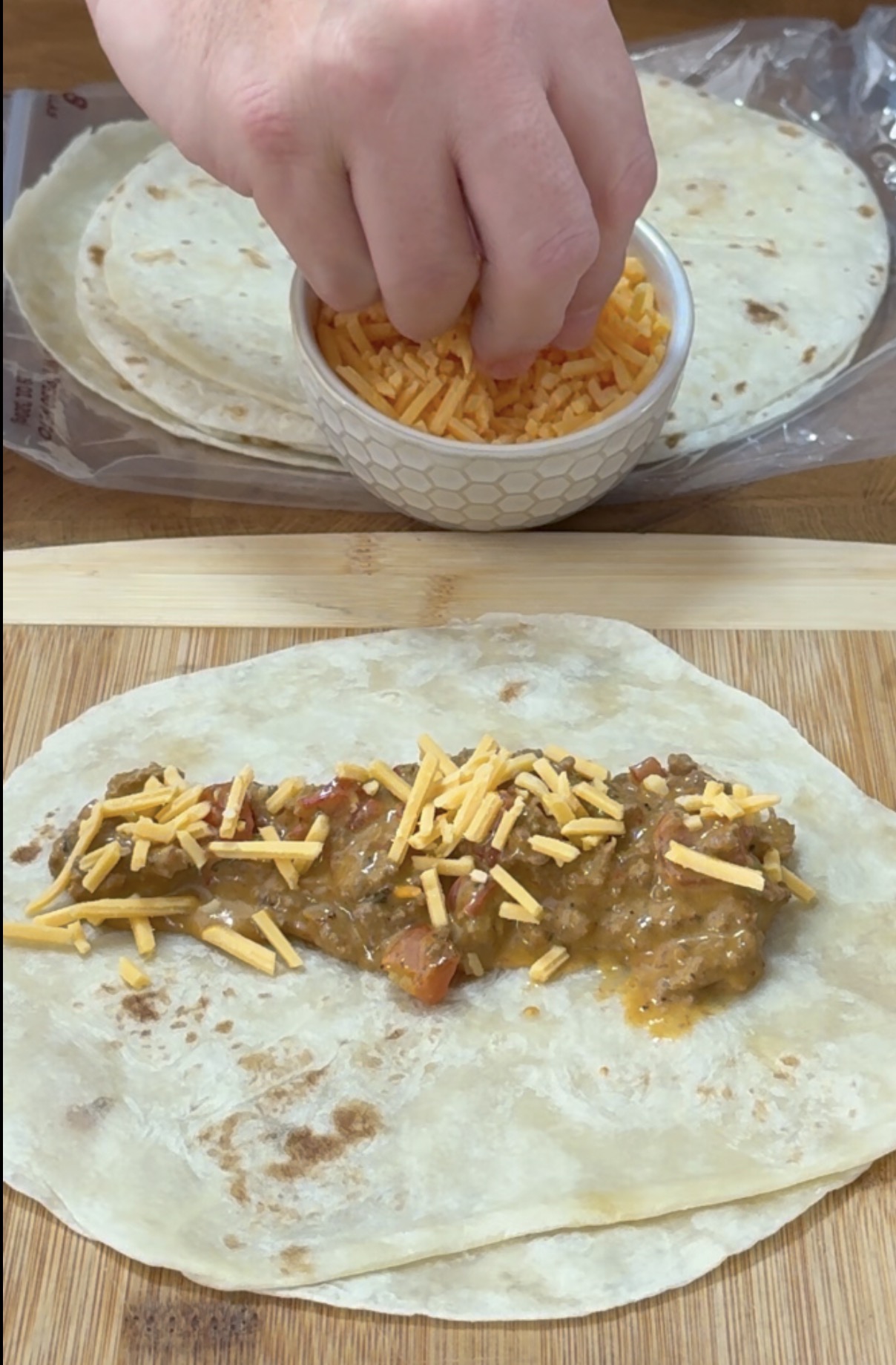 A hand sprinkles shredded cheese onto a scoop of meat filling placed on a flour tortilla, with a bowl of cheese and a stack of tortillas nearby.