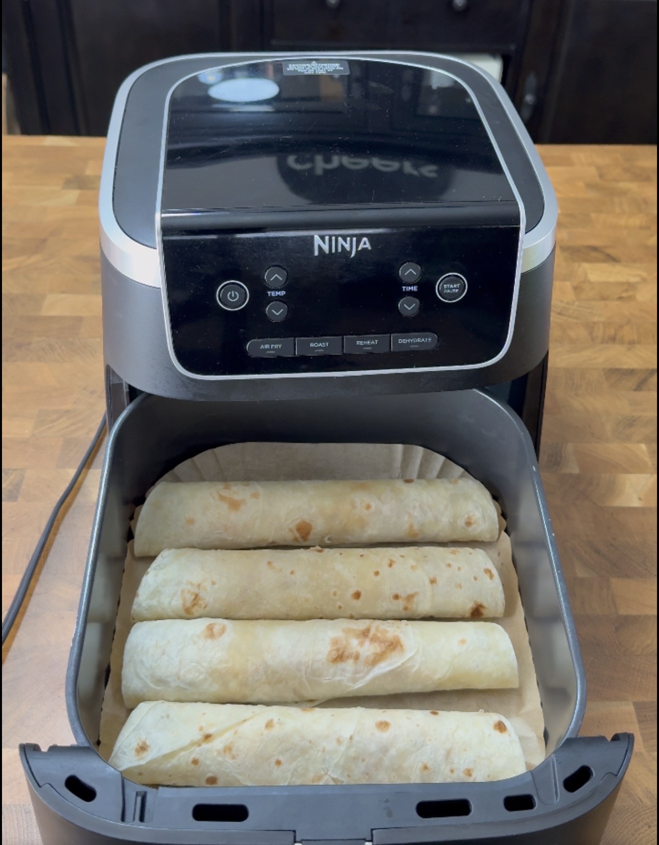 A Ninja air fryer with its basket pulled out, containing four flour tortillas rolled up on a piece of parchment paper.