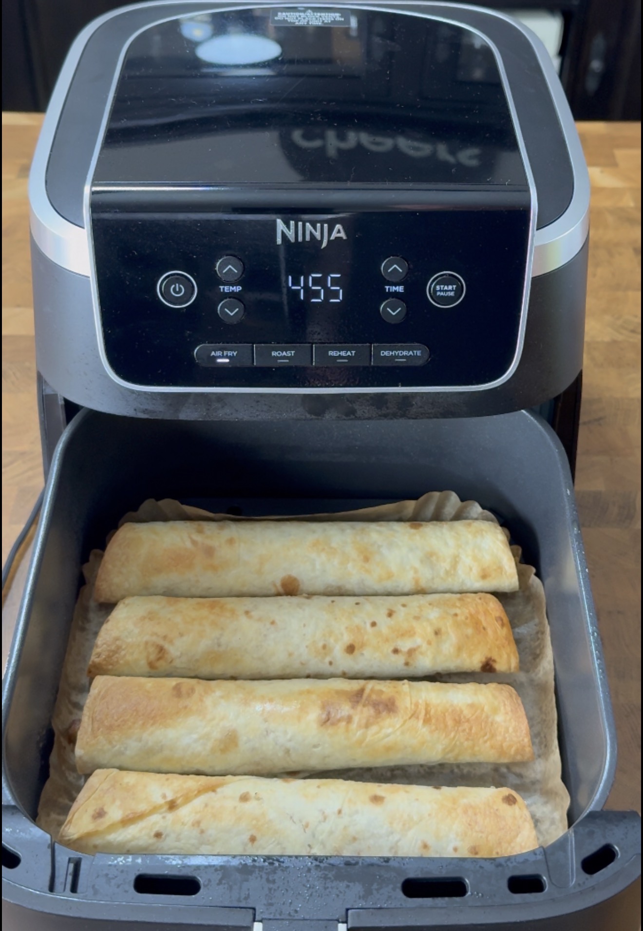 Four cylindrical wraps arranged side-by-side in the open basket of a Ninja air fryer set to 455 degrees.