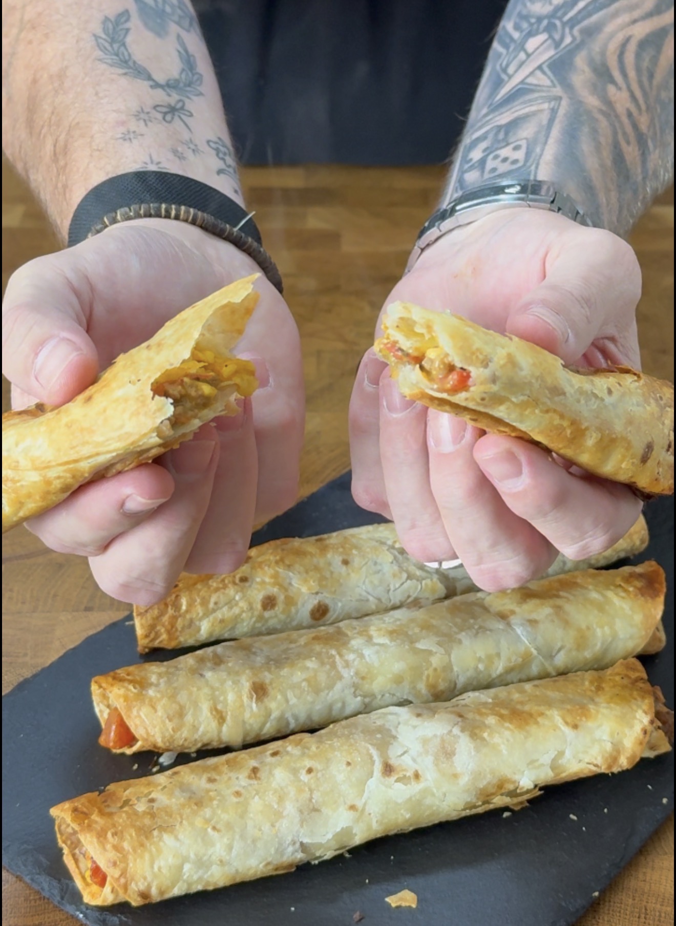 Hands pull apart a crispy, filled pastry roll, with several more whole rolls resting on a dark board below.