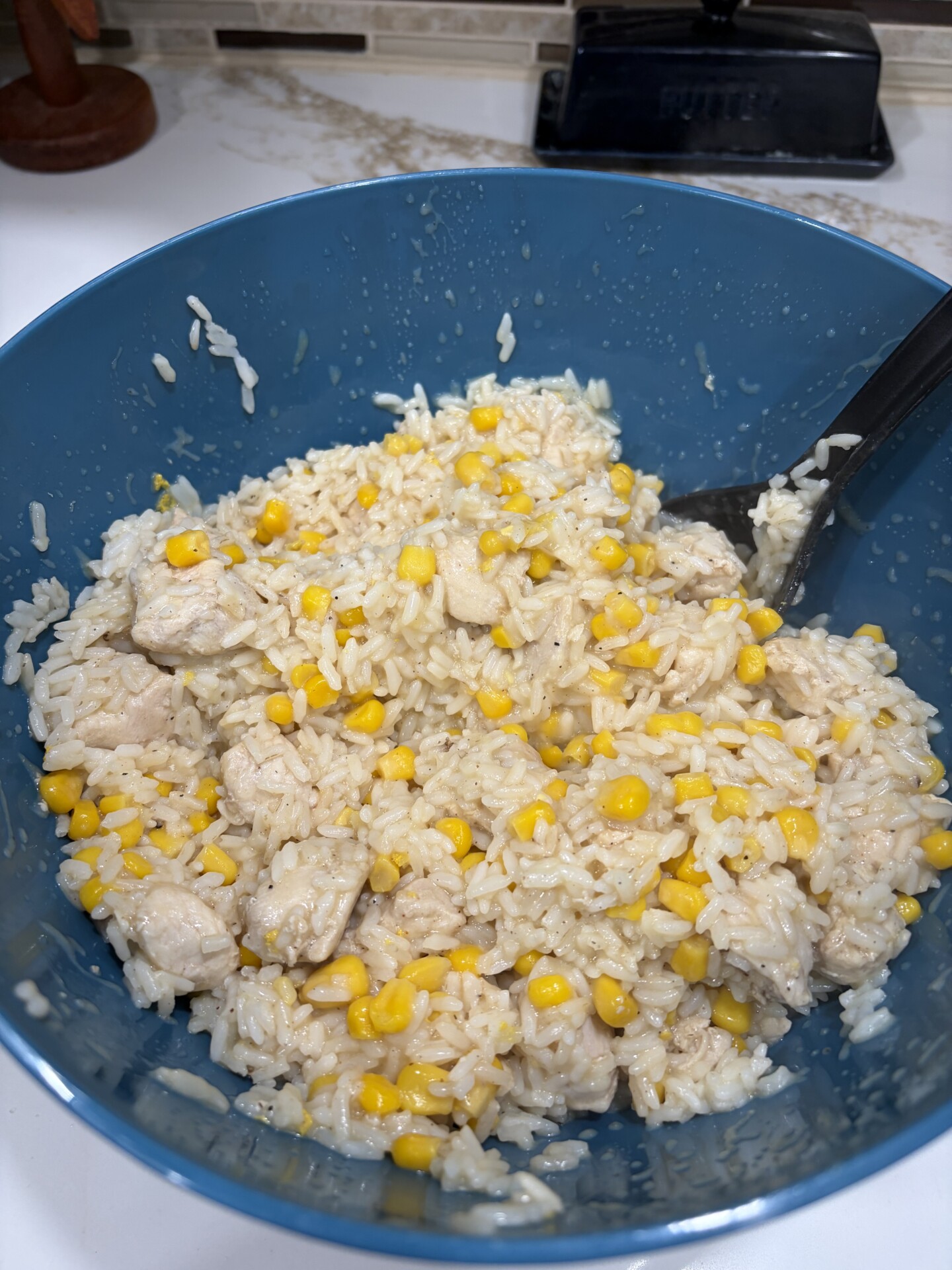 A blue bowl filled with cooked white rice mixed with yellow corn and pieces of chicken, with a black serving spoon inside.