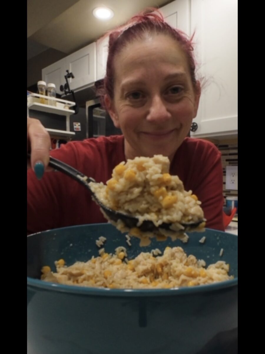A woman with pink hair smiles at the camera while holding a large spoonful of rice and corn over a teal bowl in a kitchen.