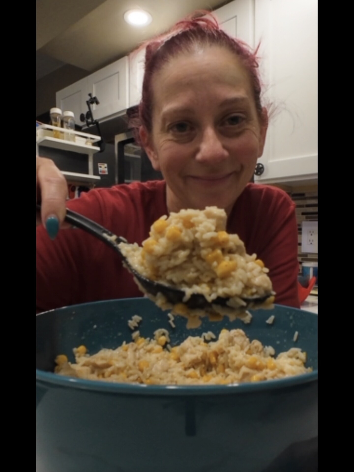 A woman with pink hair smiles at the camera while holding a large spoonful of rice and corn over a teal bowl in a kitchen.