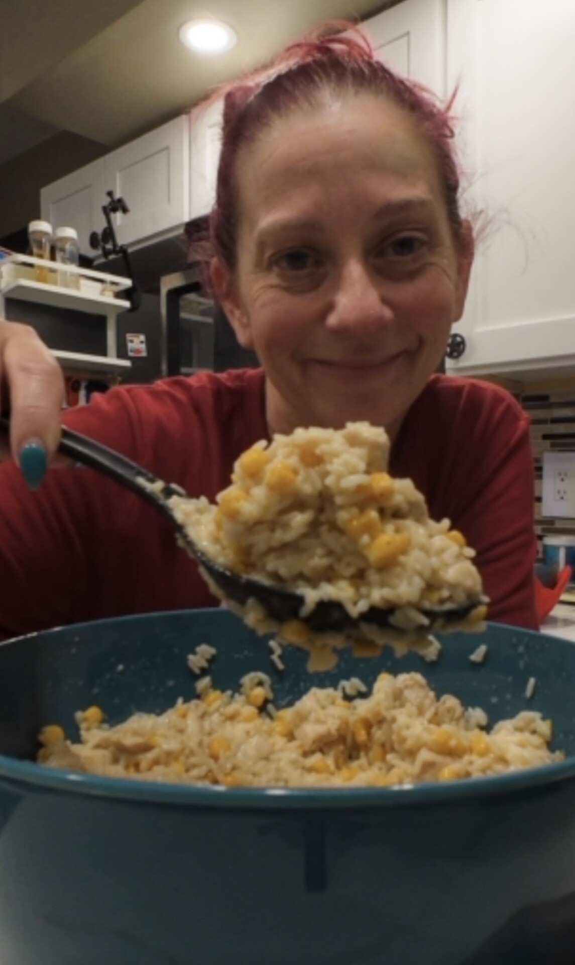 A smiling person with red hair holds up a spoonful of rice and corn from a large blue bowl in a kitchen, ready to serve the food.