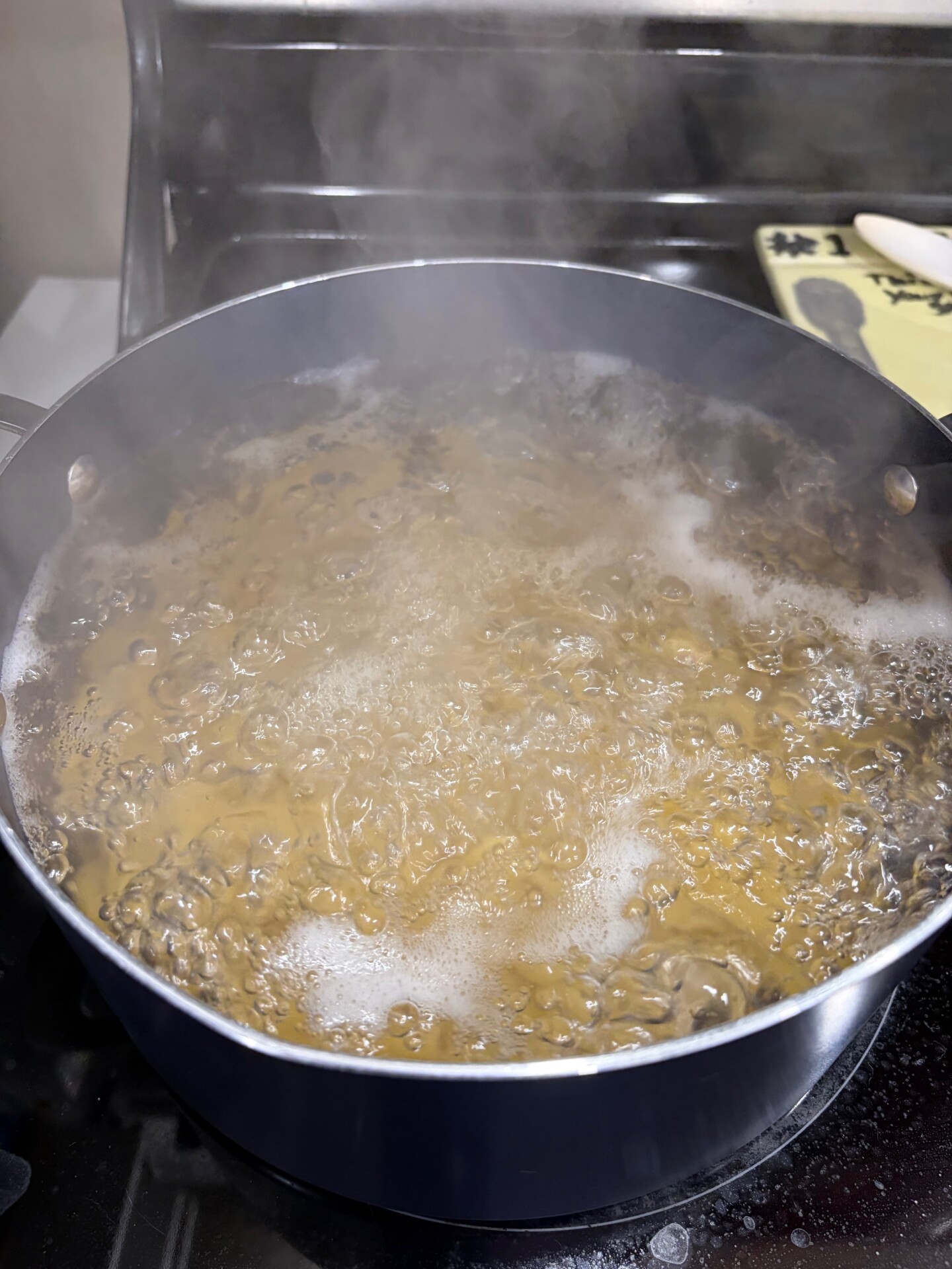 A pot of water is boiling on a stovetop, producing steam. The pot is dark-colored, and bubbles cover the waters surface. In the background, part of a yellow cutting board and a knife are visible.
