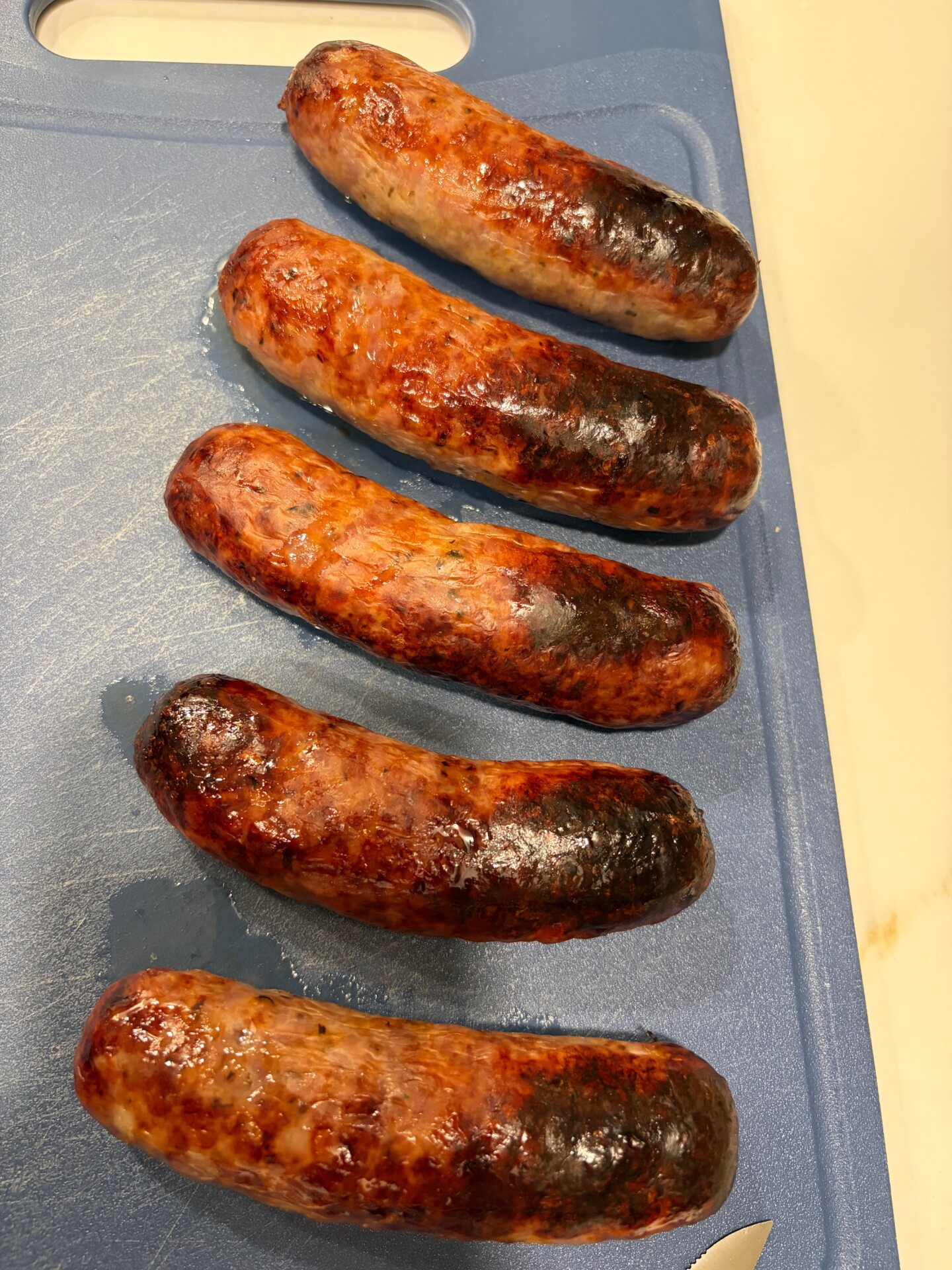 Five grilled sausages with charred spots are lined up on a blue cutting board, with part of a white countertop visible in the corner.