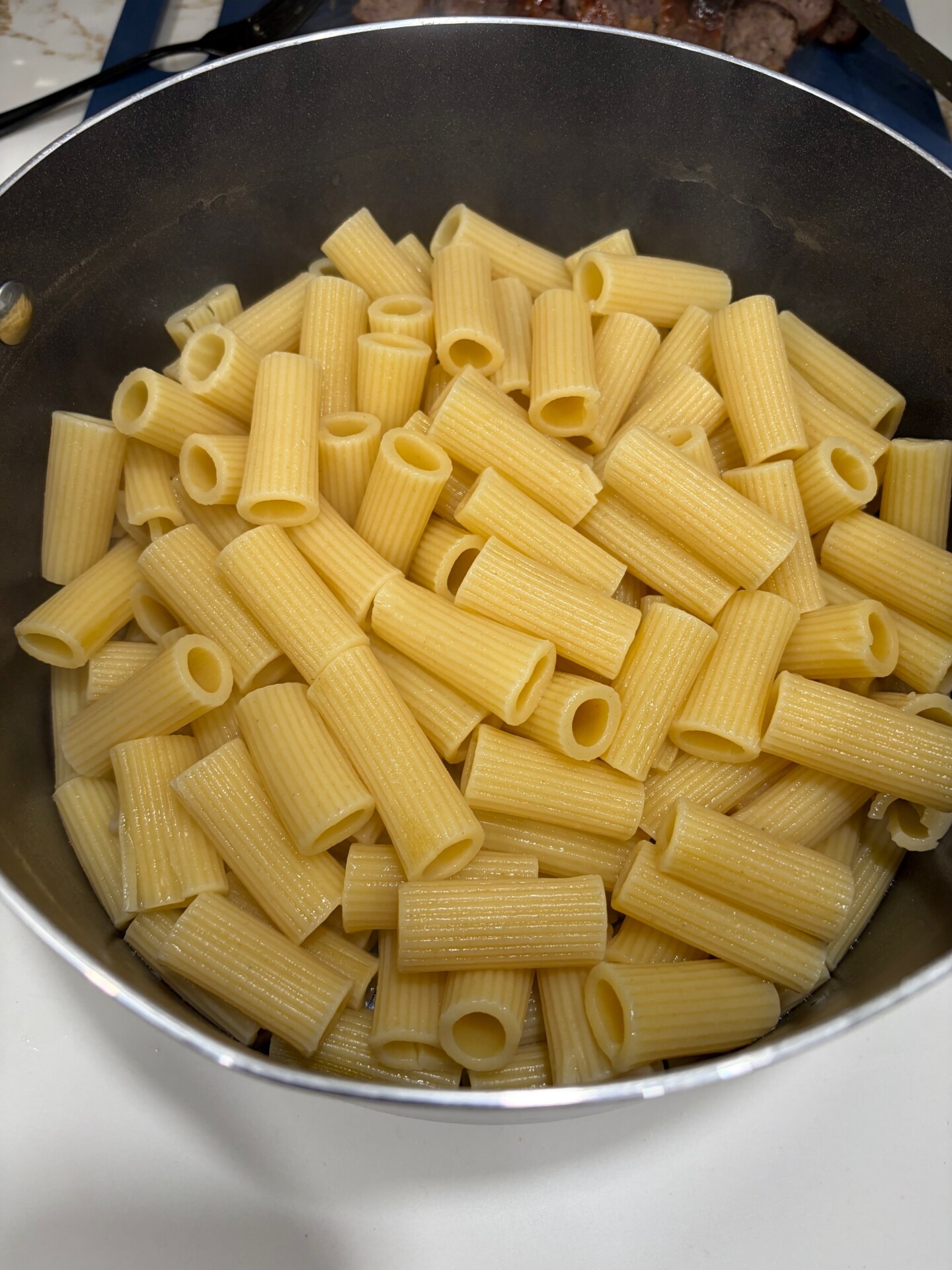 A close-up of cooked rigatoni pasta in a metal pot, showing the ridged, tube-shaped noodles.