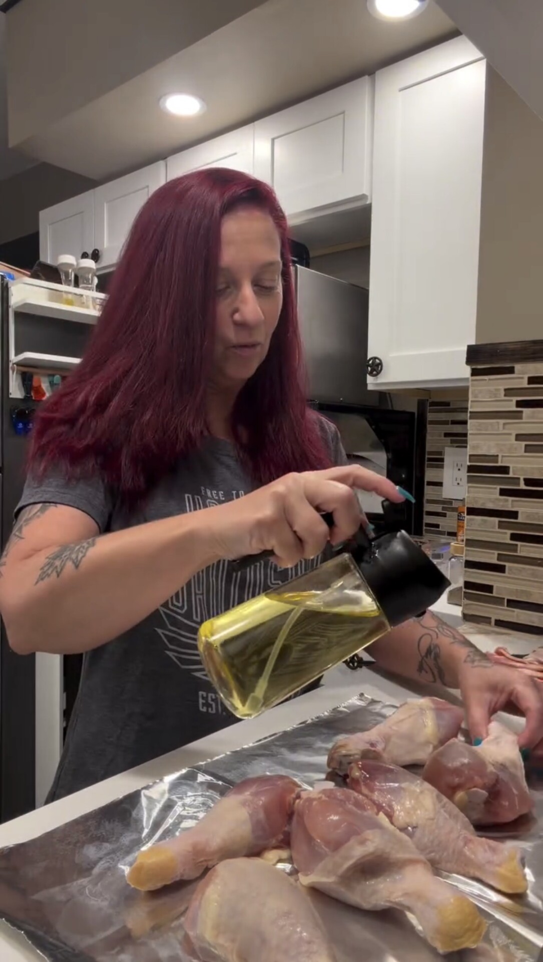 A woman with long red hair pouring oil from a glass dispenser onto raw chicken drumsticks arranged on a foil-lined baking sheet.