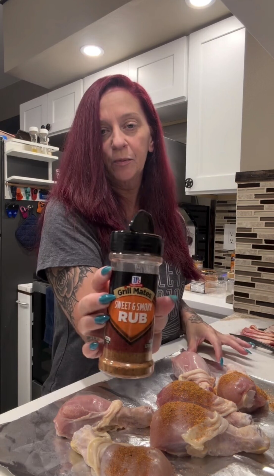 A woman holds up a bottle of McCormick Grill Mates Sweet & Smoky Rub over a baking sheet of raw chicken drumsticks seasoned with the same spice.