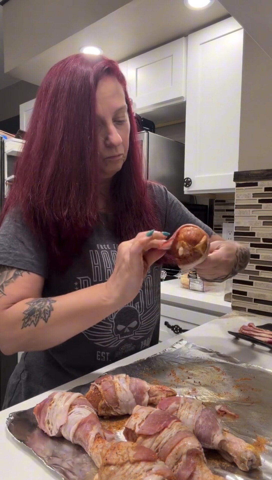 A woman wraps a strip of bacon around a seasoned chicken drumstick on a foil-lined baking sheet.