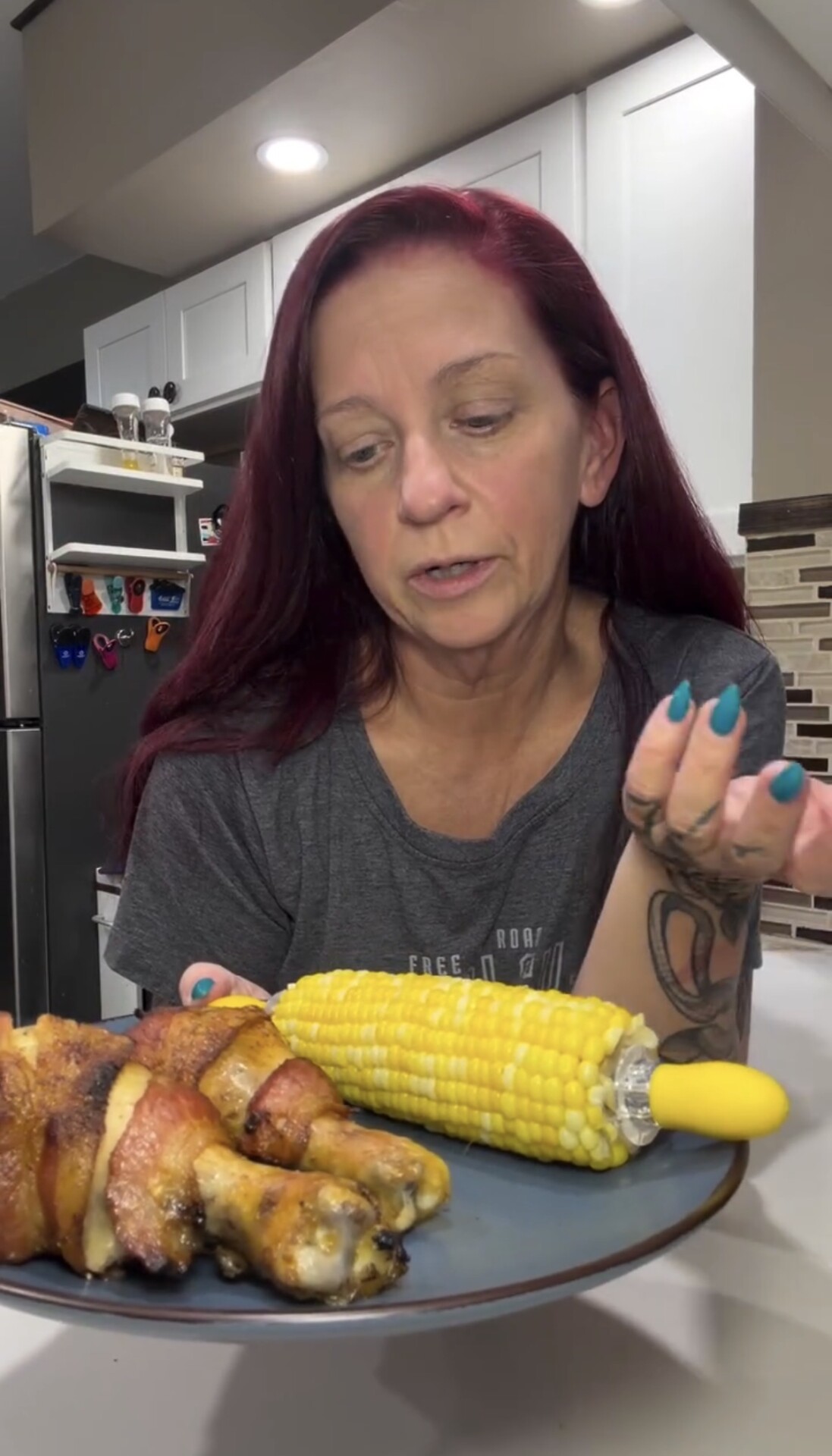 A woman holds a plate containing bacon-wrapped chicken drumsticks and an ear of corn on a yellow holder.