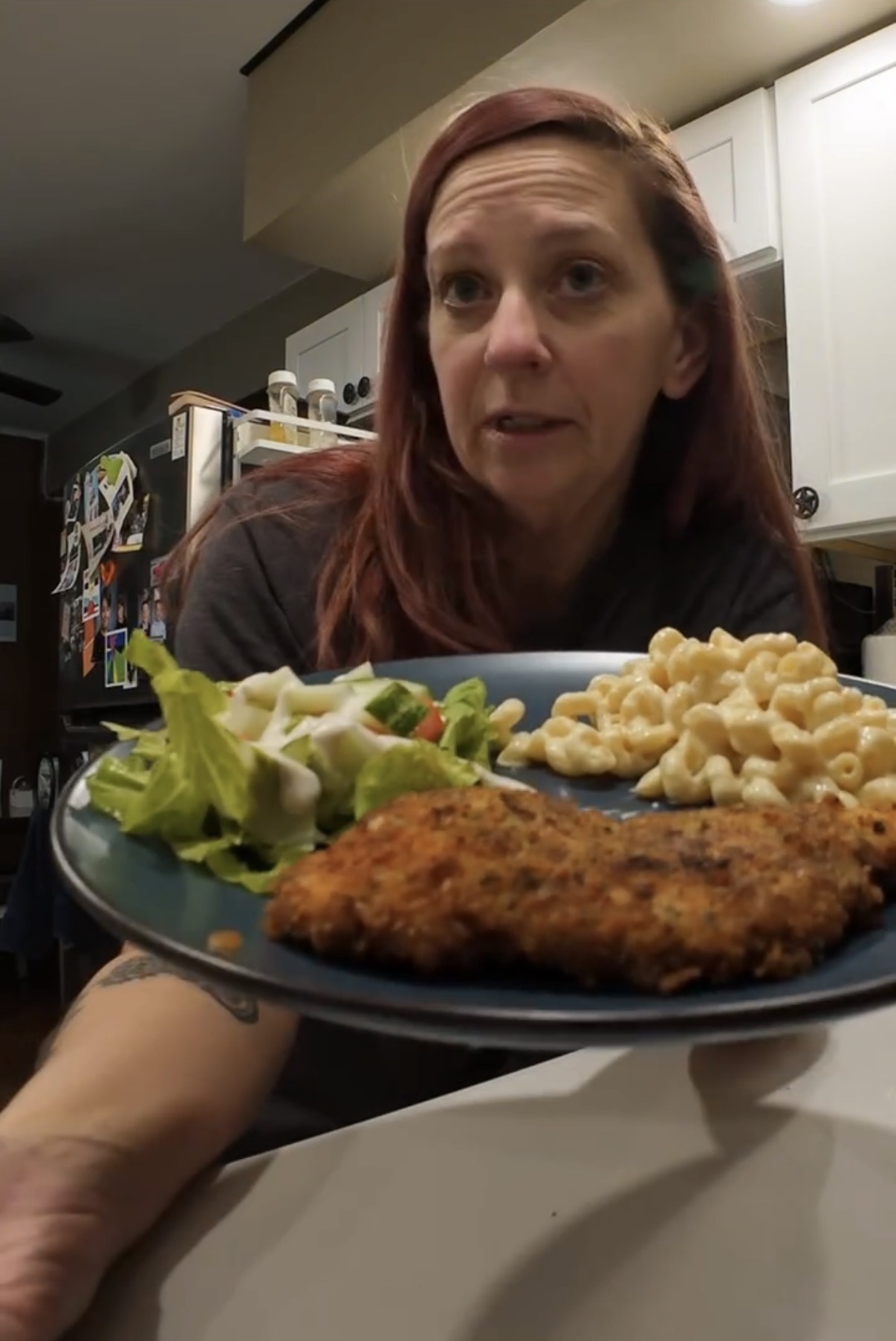 A woman holds a plate containing a breaded cutlet, macaroni and cheese, and a side salad.