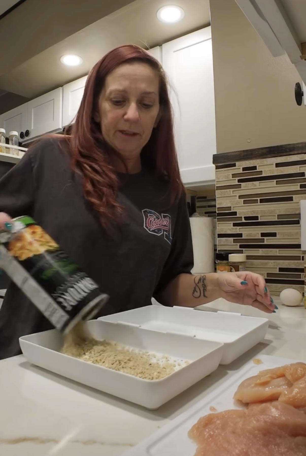 A woman pouring breadcrumbs into a white dish in a kitchen next to raw chicken breasts on a cutting board.