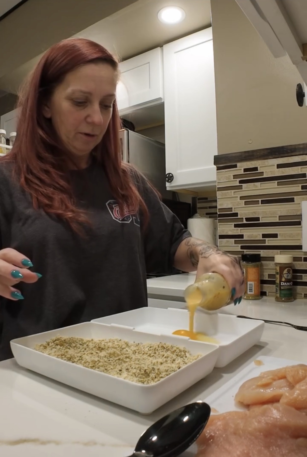 A woman pours egg wash into a shallow dish next to a dish of breadcrumbs and raw chicken breasts on a kitchen counter.