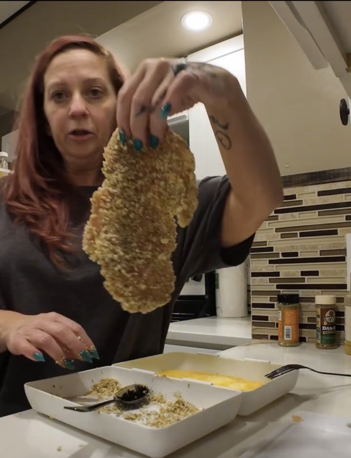 A person holds a piece of breaded, uncooked chicken above a shallow tray of breadcrumbs and a bowl of egg wash.