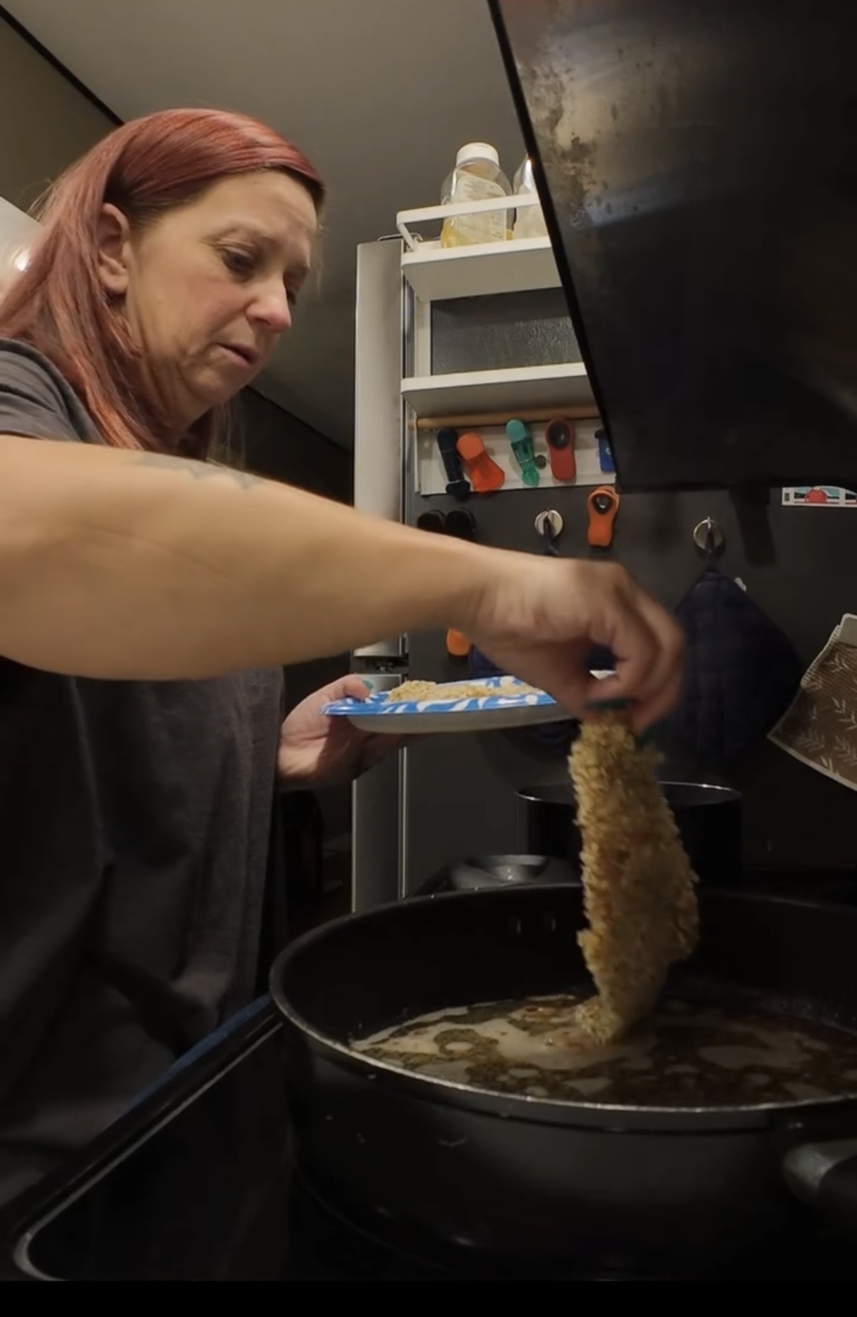 A person with reddish hair places a breaded cutlet into a pan of hot oil.
